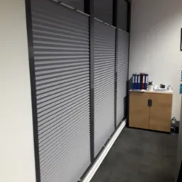 Szare, poziome osłony okienne zamontowane w biurze, widoczne z boku, z szafką na dokumenty w tle.