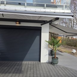 Hometechnic Sławomir Ruszczyk - Zewnętrzna roleta w kolorze antracytu i rozłożona markiza nad tarasem z desek kompozytowych. Dwa kwiaty w donicach. Widok na ogród.