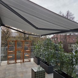Hometechnic Sławomir Ruszczyk - Szara markiza tarasowa z czarną konstrukcją na balkonie z roślinami w donicach i drewnianą konstrukcją. Widok na budynki w tle.