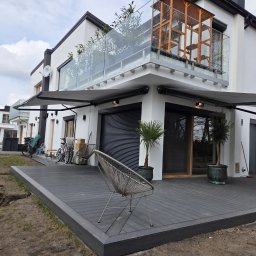 Hometechnic Sławomir Ruszczyk - Nowoczesny dom z szarą markizą nad tarasem i balkonem z szklaną balustradą. Widoczny fragment tarasu z desek kompozytowych i przygotowany teren pod trawnik.