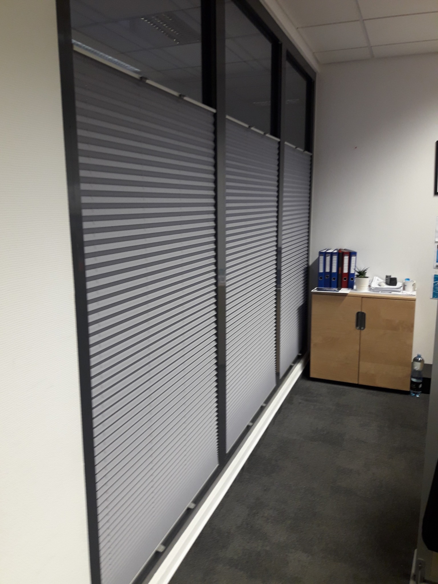 Szare, poziome osłony okienne zamontowane w biurze, widoczne z boku, z szafką na dokumenty w tle.