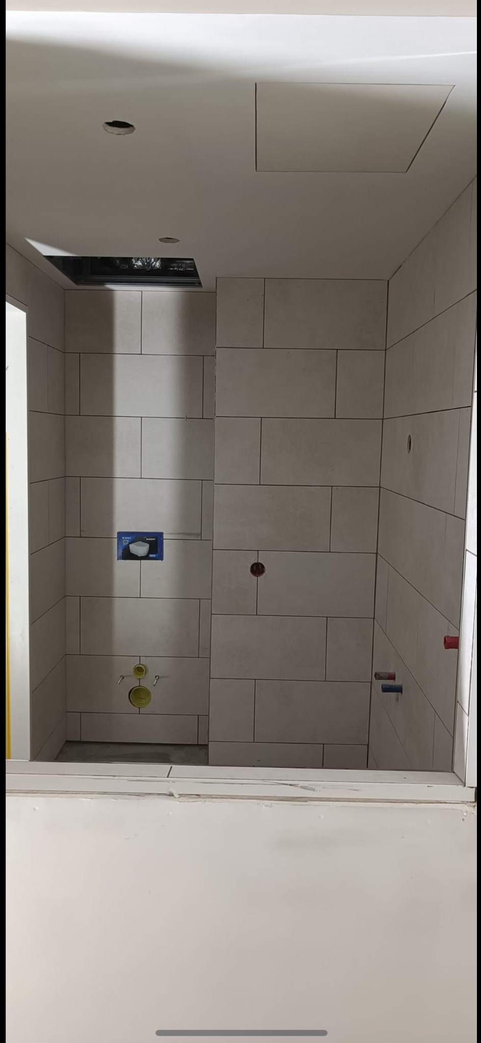 Świeżo wykafelkowana ściana kabiny prysznicowej z widocznymi przyłączami wodnymi i odpływowymi w trakcie montażu armatury, z widoczną instalacją podtynkową.