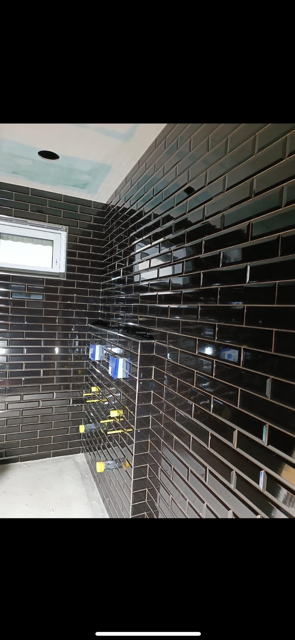 Ściana wyłożona błyszczącymi, ciemnymi, prostokątnymi płytkami ceramicznymi przypominającymi cegiełki, z widocznymi elementami instalacji wodnej i poziomnicami, w trakcie prac wykończeniowych...