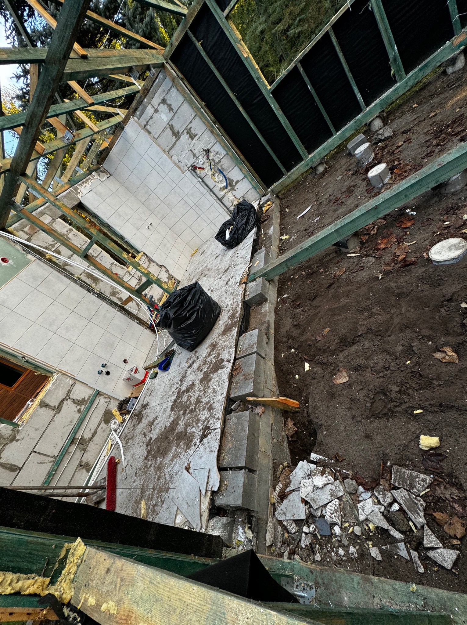 Widok z góry na plac budowy z elementami rozbiórki i przygotowania do budowy, widoczne fragmenty ścian z białymi płytkami, drewniana konstrukcja dachu, ziemia, gruz, dwa czarne worki na śmieci...