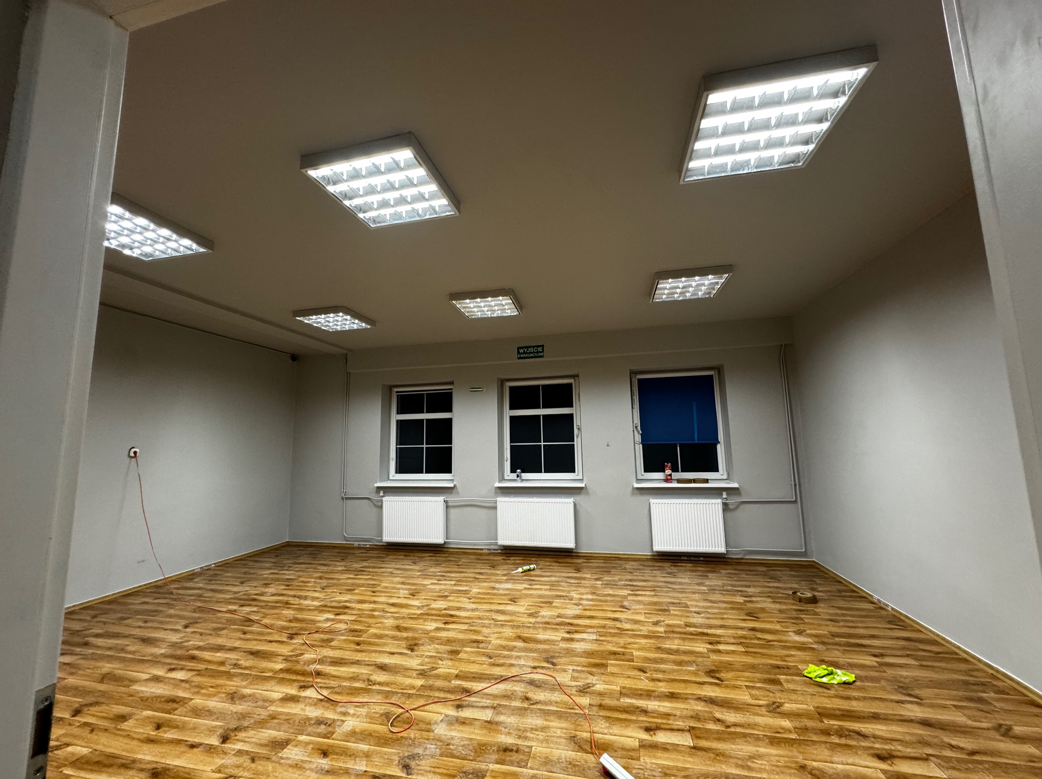 Pusty pokój po remoncie z panelami podłogowymi w odcieniach brązu, trzema oknami z grzejnikami i białymi ścianami oraz sześcioma lampami sufitowymi.