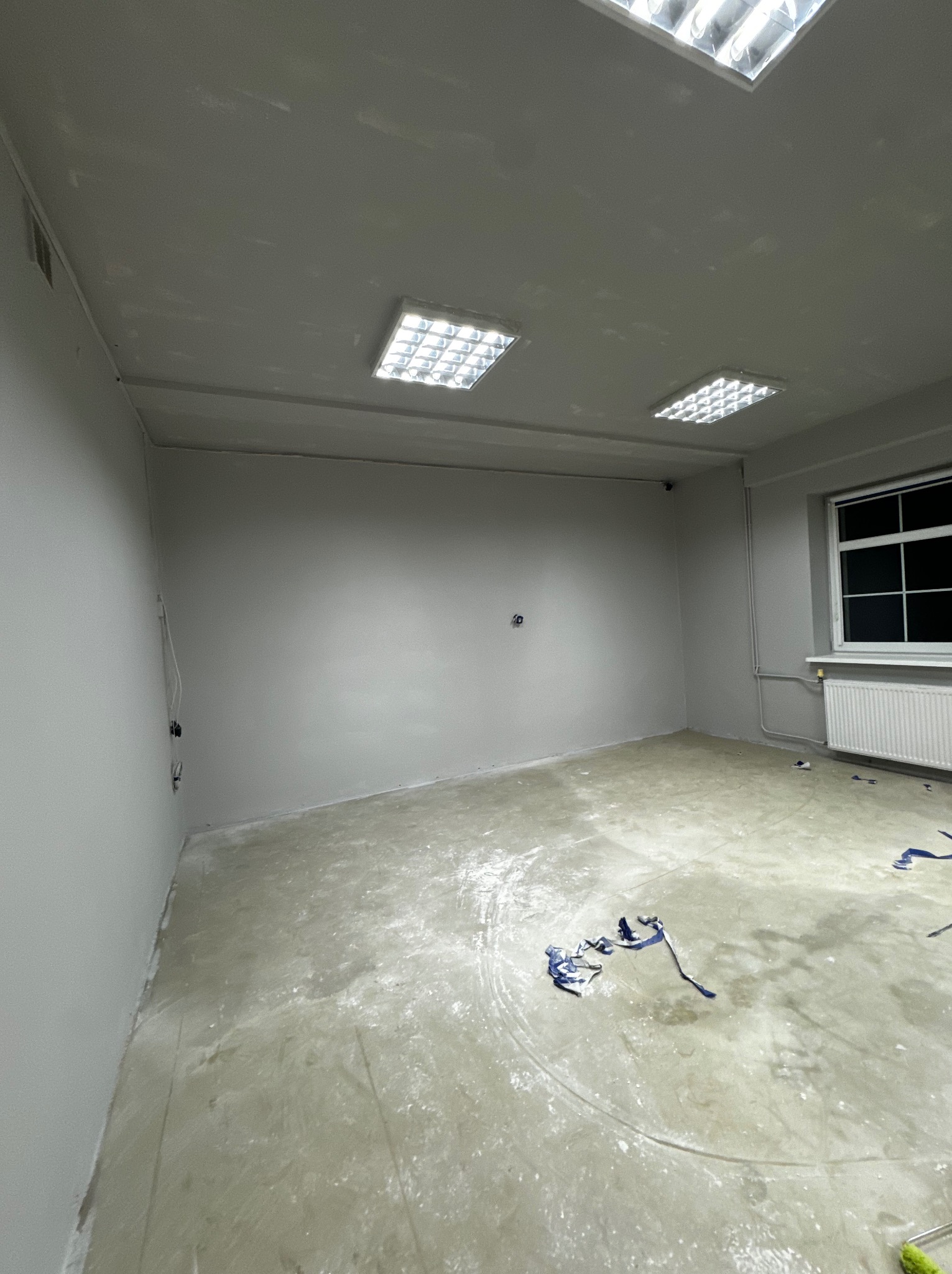 Szare, puste pomieszczenie po malowaniu ścian, widoczne ślady farby na podłodze i taśmy malarskie, okno z grzejnikiem, cztery lampy sufitowe.