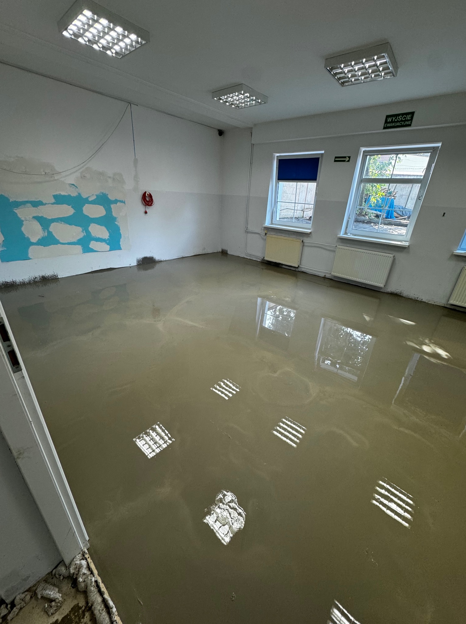 Wylewka samopoziomująca na podłodze w pomieszczeniu z widocznymi grzejnikami, dwoma oknami i śladami po usuniętej glazurze na ścianie.