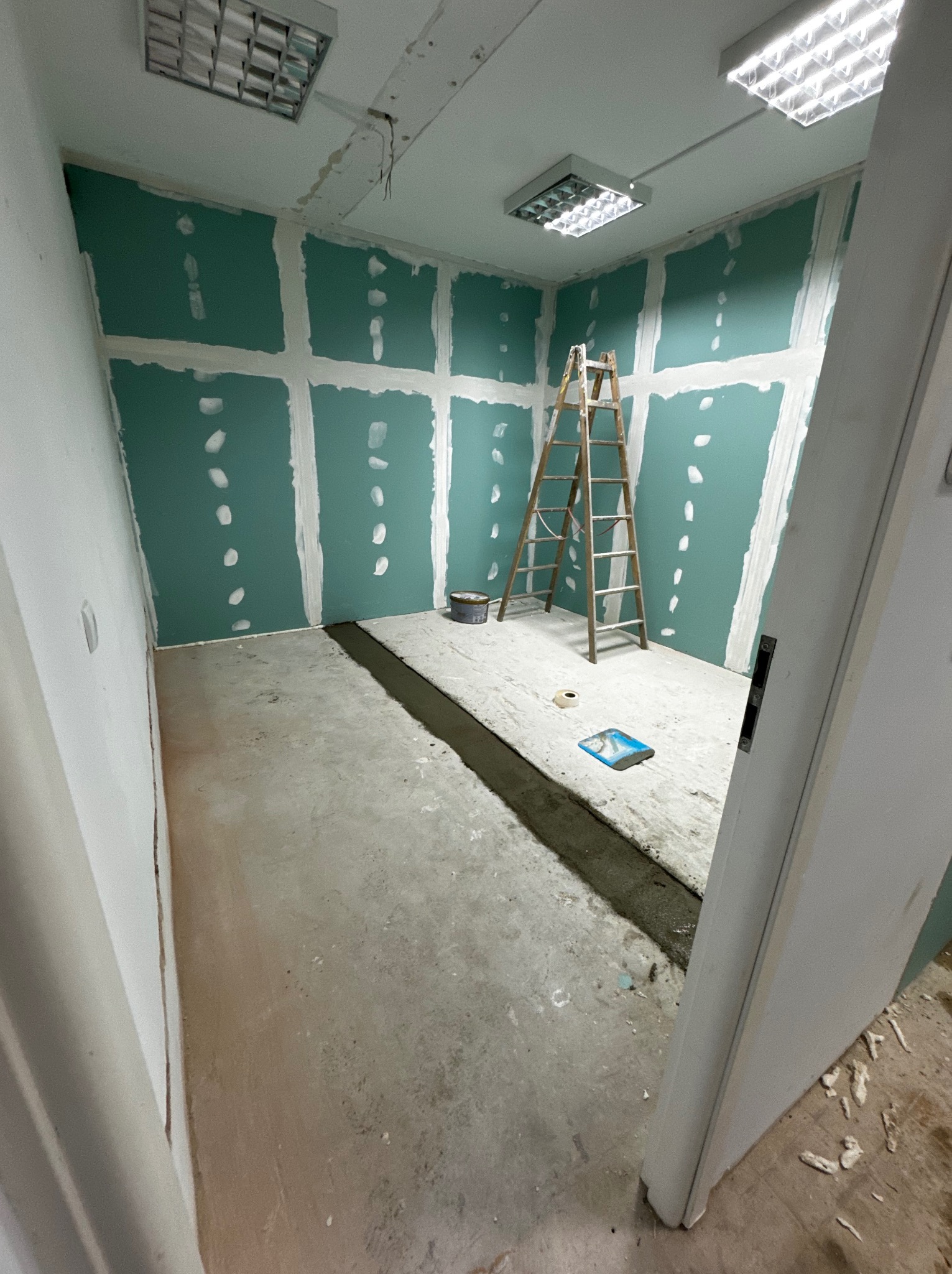 Wnętrze pokoju w trakcie remontu: zielone płyty karton-gips na ścianach z widocznymi łączeniami, drabina drewniana, szpachla w wiaderku, listwa podłogowa zalana betonem, surowy beton na podłodze...