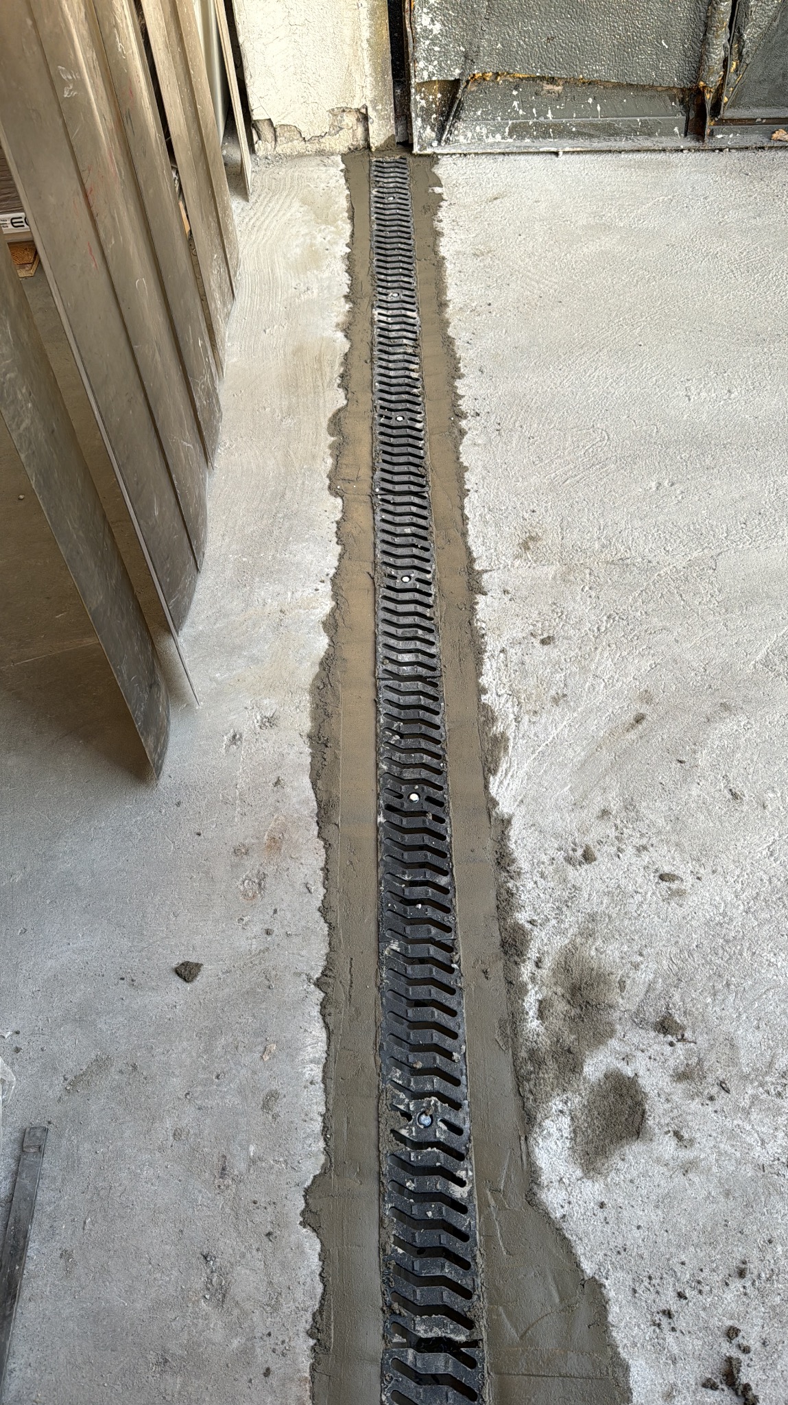 Montaż liniowego odpływu podłogowego z czarną kratką w szarej posadzce betonowej, widoczne ślady świeżego cementu i narzędzia budowlane.