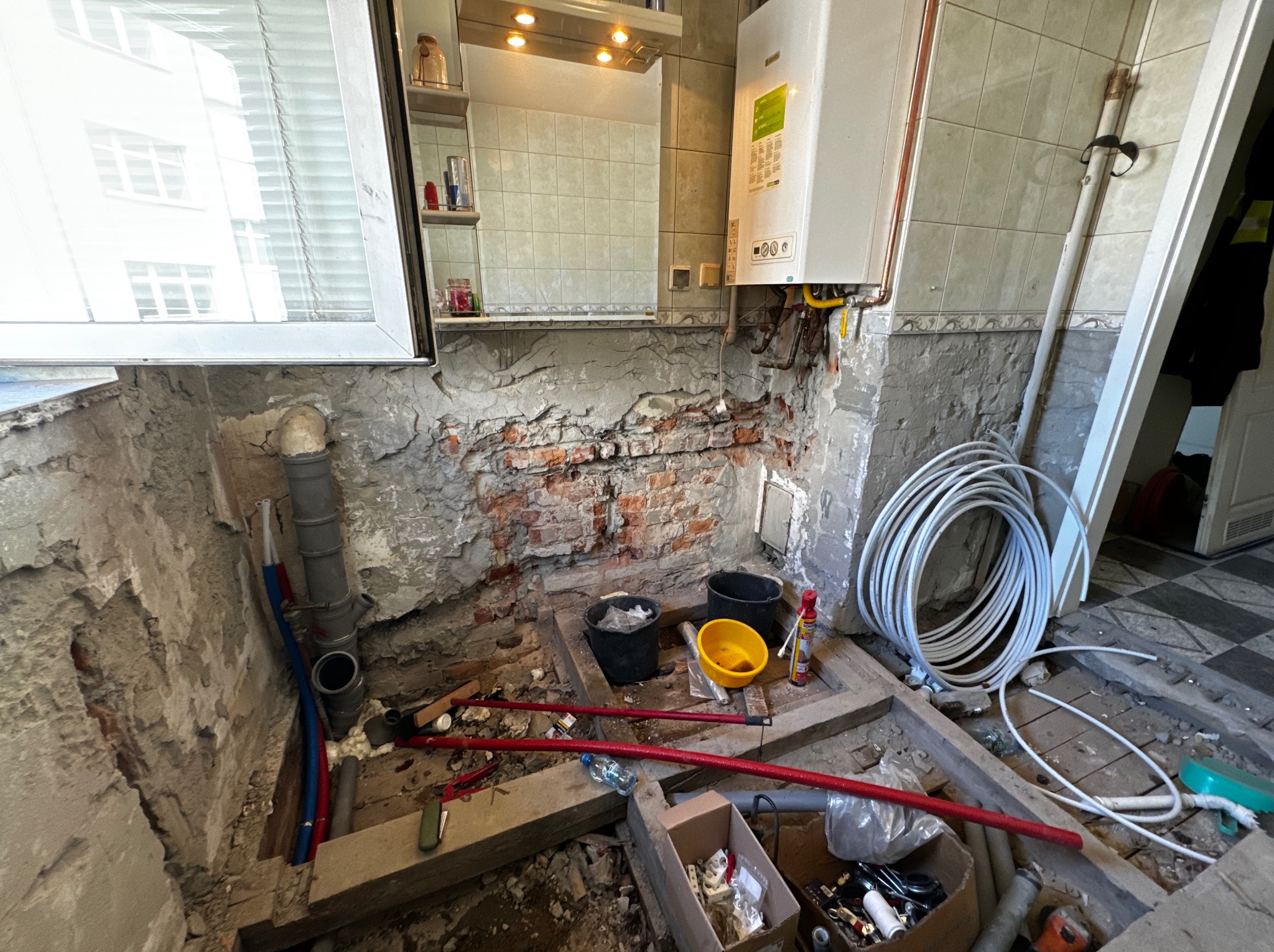 Niedokończona łazienka w trakcie demontażu: odsłonięta ceglana ściana, stare rury, wiadro, narzędzia, zwój białych rur, częściowo usunięte płytki, widoczny fragment okna i lustra z szafką.