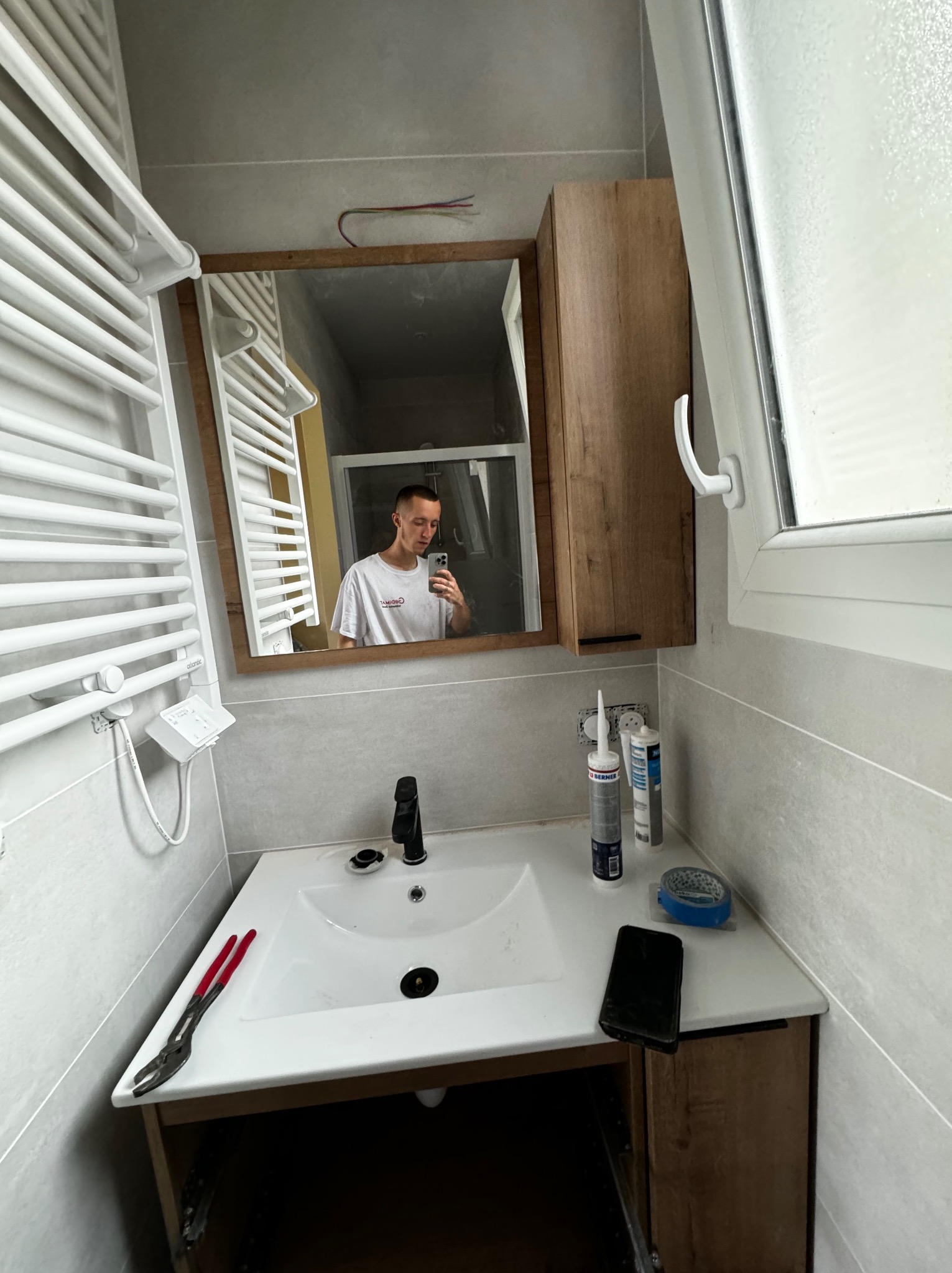 Łazienka w trakcie wykańczania: widoczny biały grzejnik drabinkowy, umywalka z czarną armaturą, silikon budowlany, narzędzia (kombinerki), oraz odbicie osoby robiącej zdjęcie w lustrze.