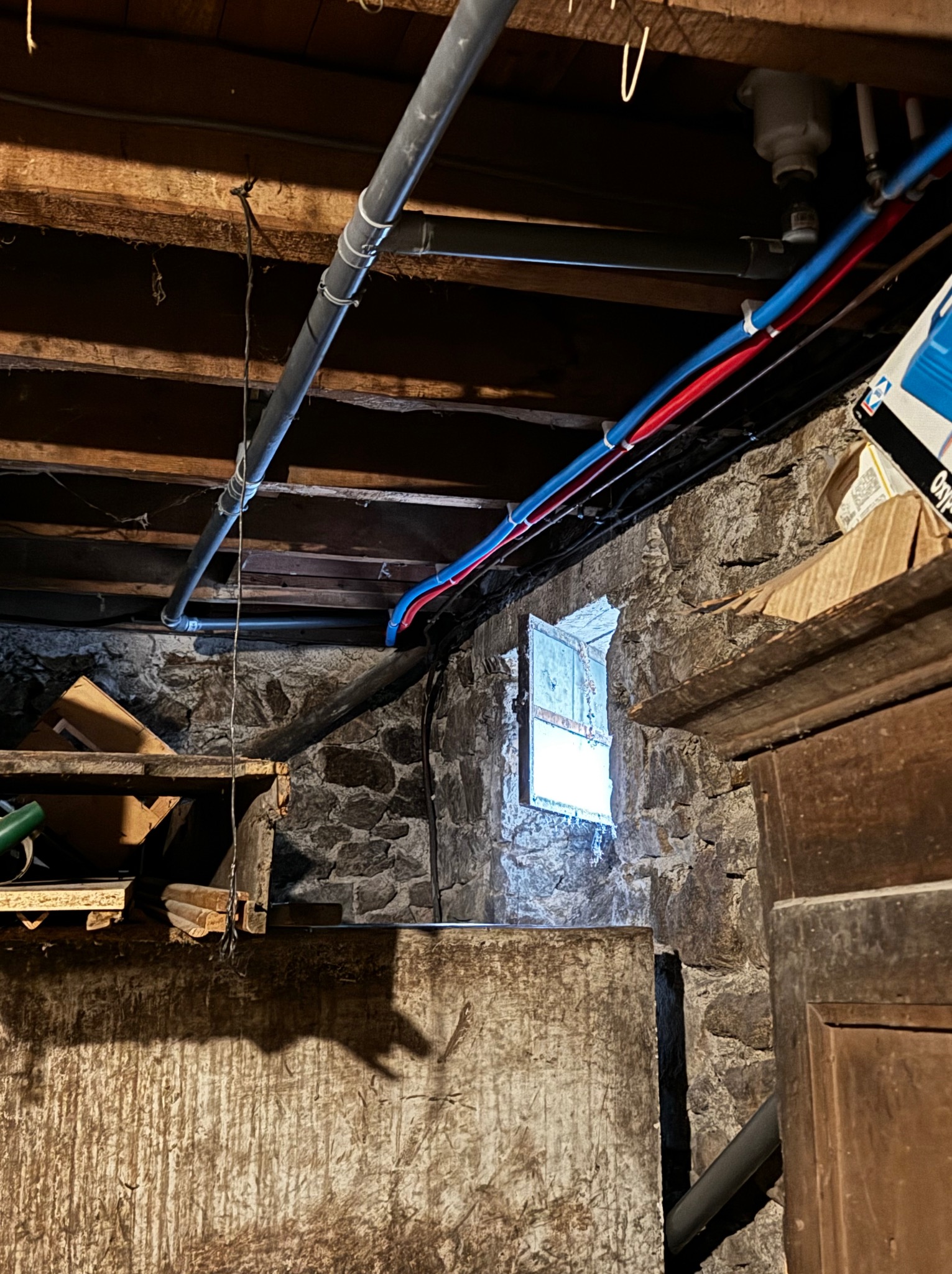 Piwnica z kamiennymi ścianami, małym oknem w głębi, drewnianym stropem i nowymi rurami instalacji wodnej w kolorach niebieskim i czerwonym.