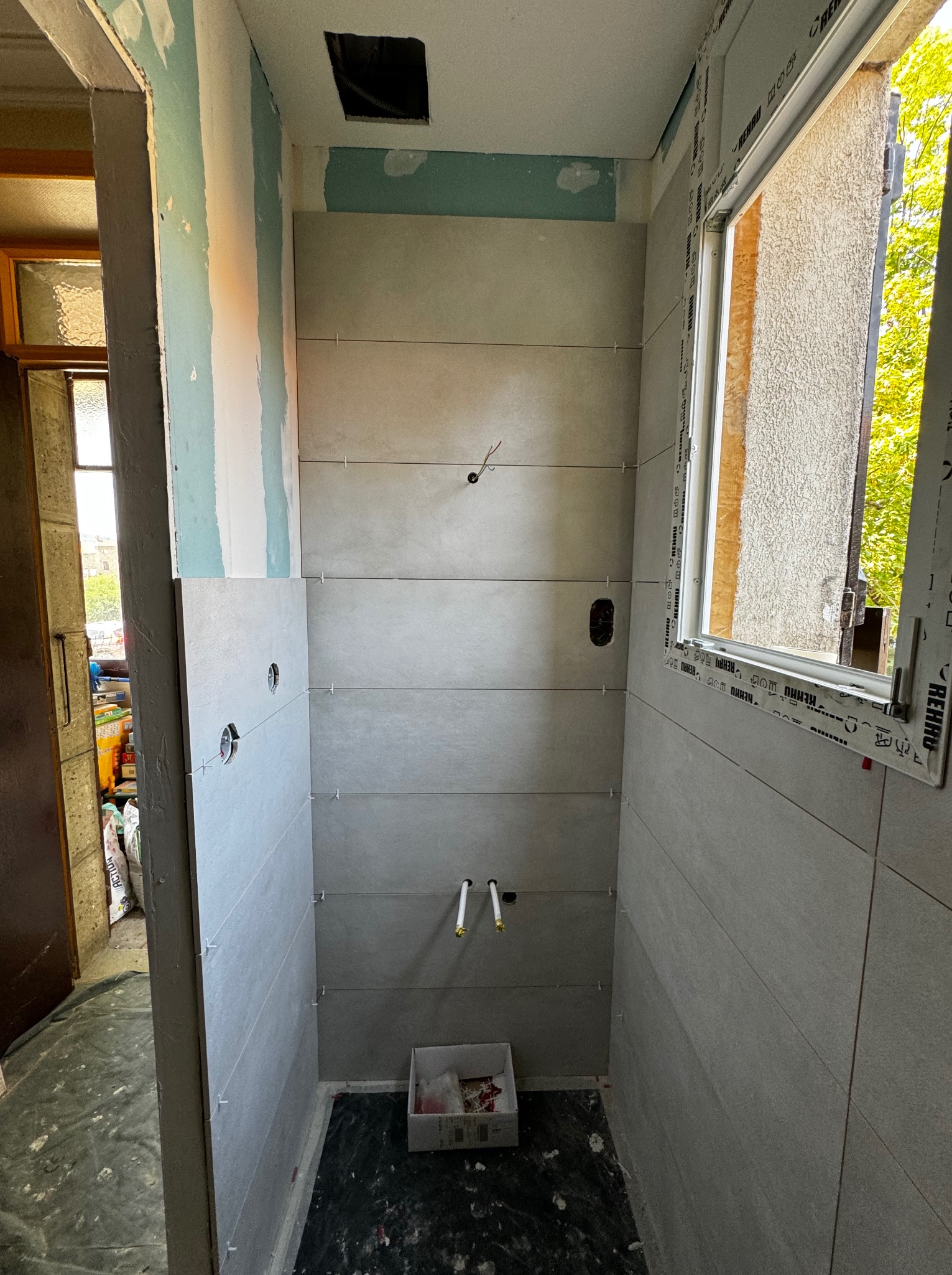 Łazienka w trakcie kładzenia płytek ceramicznych w odcieniach szarości, widoczne otwory na gniazdka elektryczne i rury, krzyżyki dystansowe między płytkami, okno z widokiem na zieleń, surowy stan...