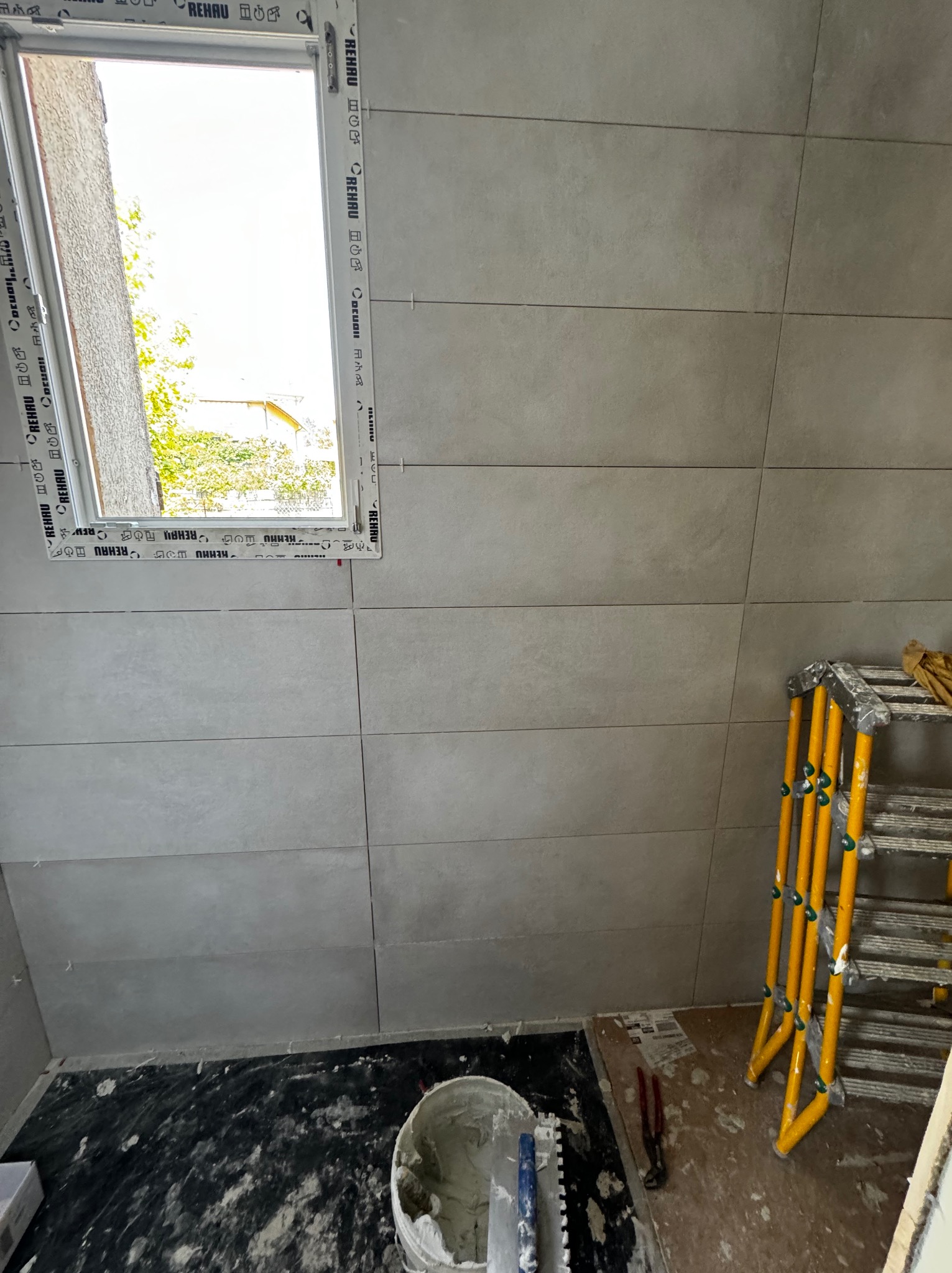 Ściana łazienki w trakcie układania szarych, podłużnych płytek ceramicznych, widoczne okno z profilem Rehau, żółta drabina, wiadro z zaprawą klejową i narzędzia glazurnicze na podłodze.