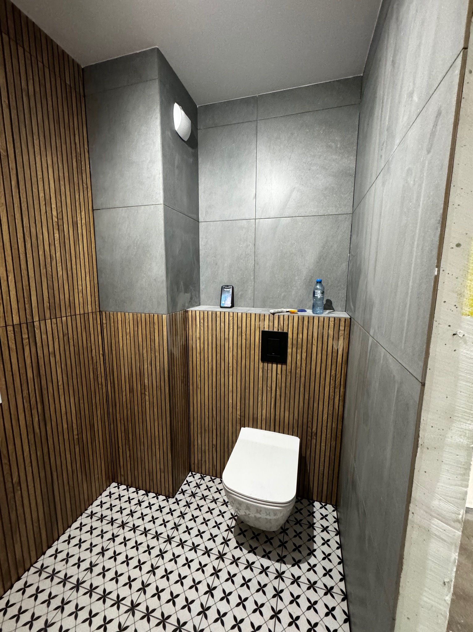 Nowoczesna łazienka w trakcie wykańczania: ściany wyłożone szarymi płytkami imitującymi beton, drewniana boazeria panelowa, wzorzysta czarno-biała podłoga, wisząca toaleta, telefon i woda na półce.