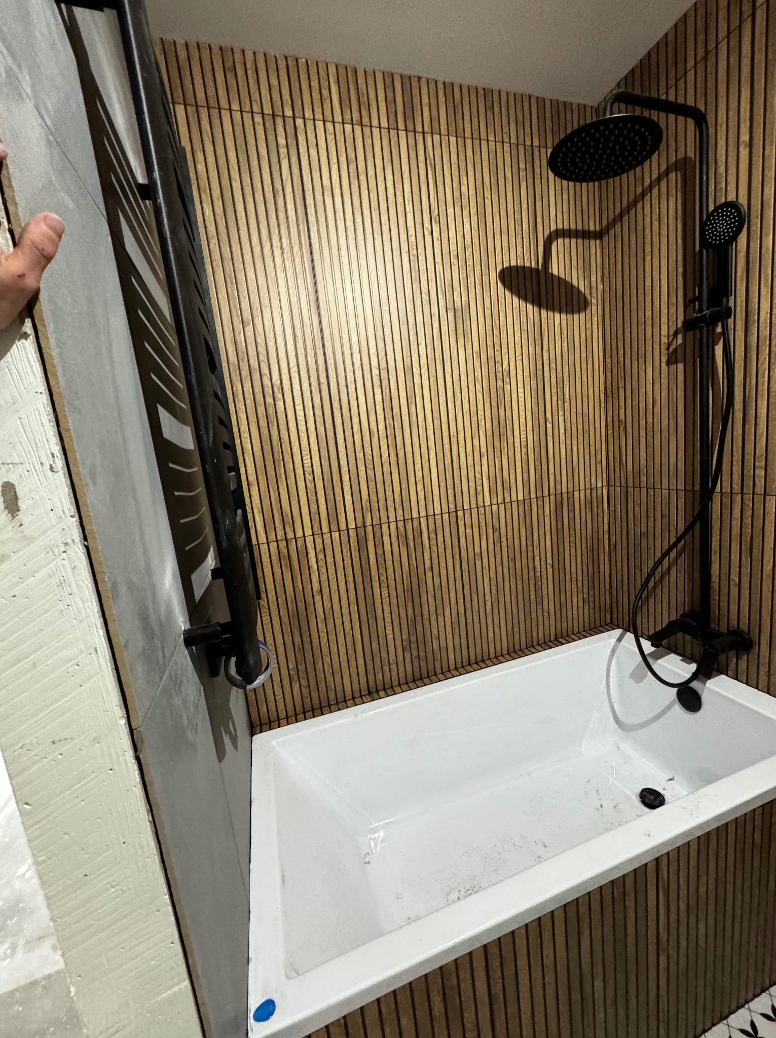 Nowoczesna łazienka w trakcie wykańczania: biała wanna, czarna armatura i drewnopodobne panele ścienne. Widoczny fragment grzejnika drabinkowego i niedokończona ściana z szarych płyt.