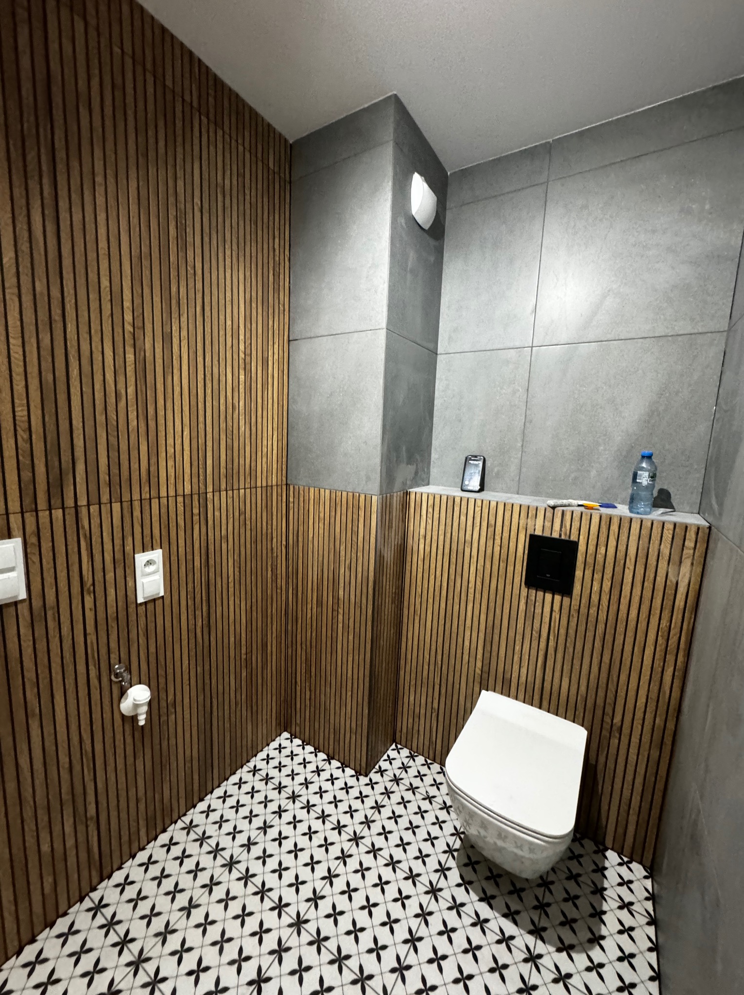 Nowoczesna łazienka z drewnianymi lamelami na ścianach, szarą glazurą, wzorzystą podłogą i podwieszaną toaletą.