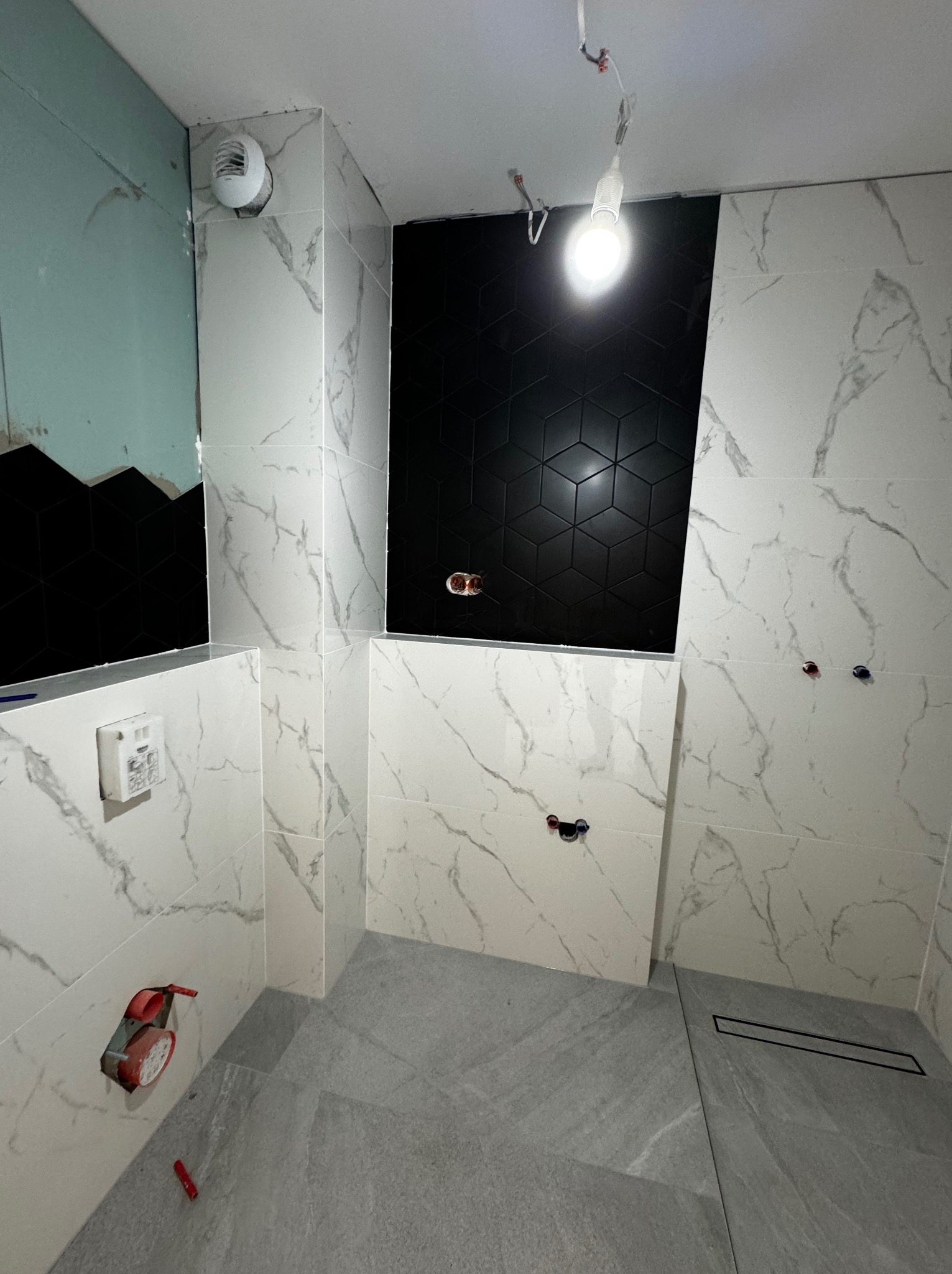 Wnętrze łazienki w trakcie wykańczania: białe płytki imitujące marmur na ścianach, fragment ściany z czarnymi, heksagonalnymi płytkami, szara podłoga z odpływem liniowym, widoczne instalacje wodne...