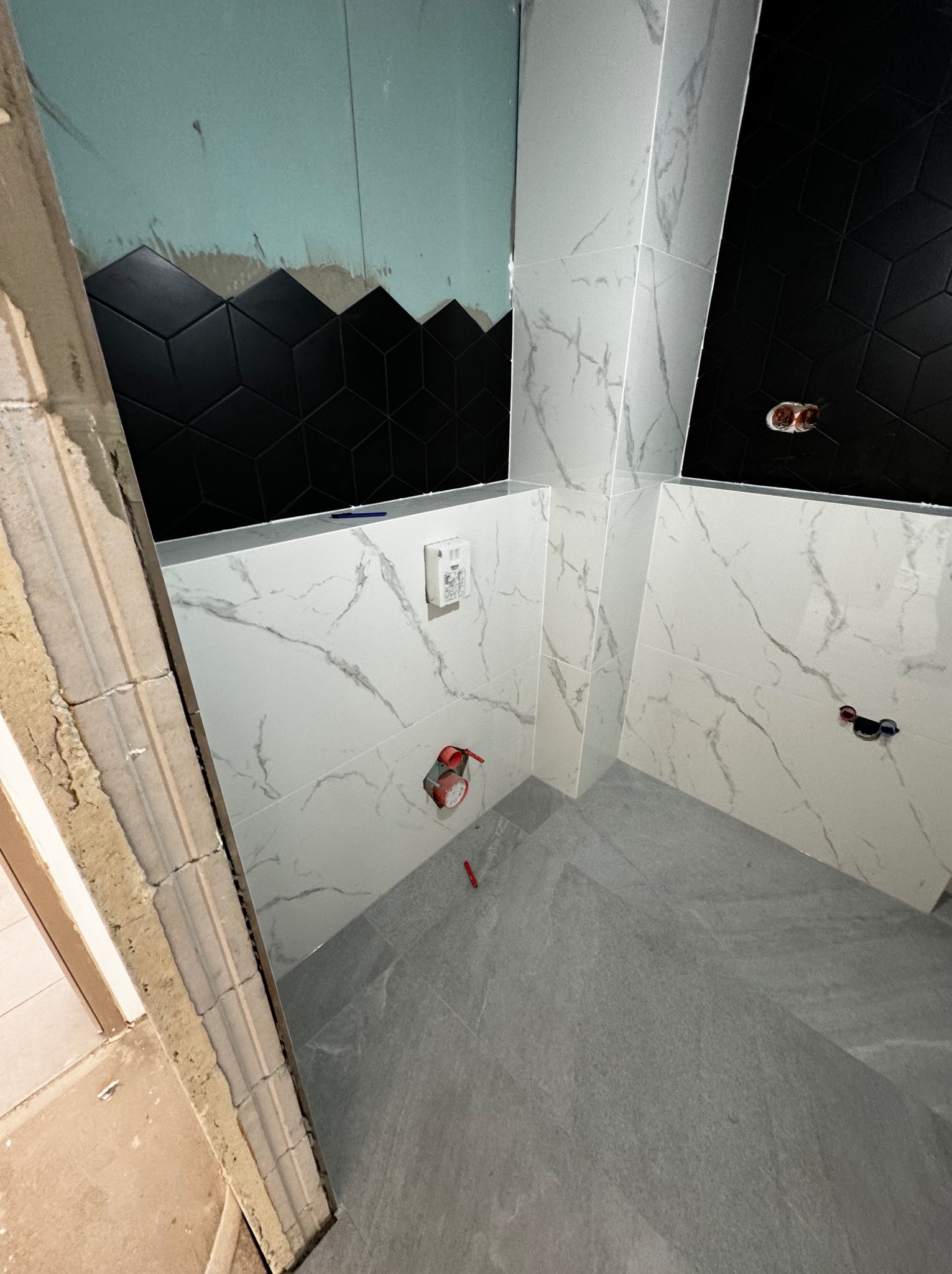 Łazienka w trakcie wykańczania: ściany wyłożone białymi płytkami imitującymi marmur oraz czarnymi płytkami o geometrycznym wzorze, widoczne instalacje hydrauliczne, surowa ściana z płytą gipsową.