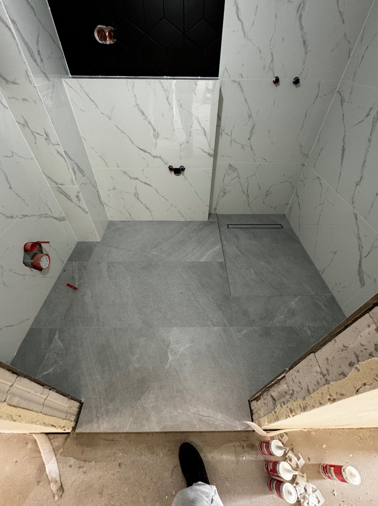 Nowoczesna łazienka w trakcie wykańczania: białe marmurowe płytki ścienne, szare płytki podłogowe z odpływem liniowym, widoczne wystające instalacje i puszki elektryczne, oraz resztki materiałów...