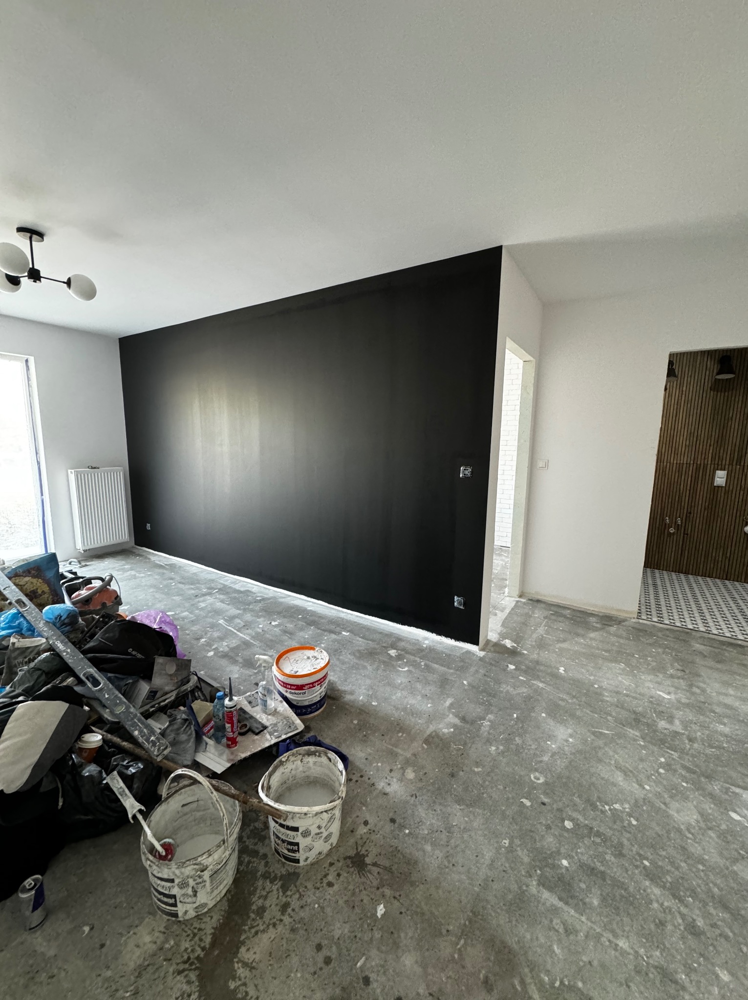Świeżo pomalowana na czarno ściana w trakcie remontu mieszkania, widoczne narzędzia malarskie, puszki z farbą, grzejnik i fragment wykończonej łazienki w tle.