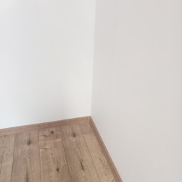 Tomasz Hejka - Jasne panele podłogowe ułożone w rogu pomieszczenia z białymi ścianami. Widoczne listwy przypodłogowe w kolorze paneli.