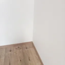 Jasne panele podłogowe ułożone w rogu pomieszczenia z białymi ścianami. Widoczne listwy przypodłogowe w kolorze paneli.