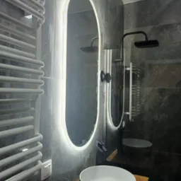 Nowoczesna łazienka z marmurowymi płytkami, podświetlanym lustrem i biało-czarną umywalką na drewnianym blacie. Grzejnik drabinkowy i czarna armatura dodają elegancji.