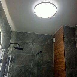 Tomasz Hejka - Nowoczesna łazienka po remoncie: ciemne płytki imitujące marmur, drewniane panele ścienne, okrągła lampa sufitowa i czarny zestaw prysznicowy.