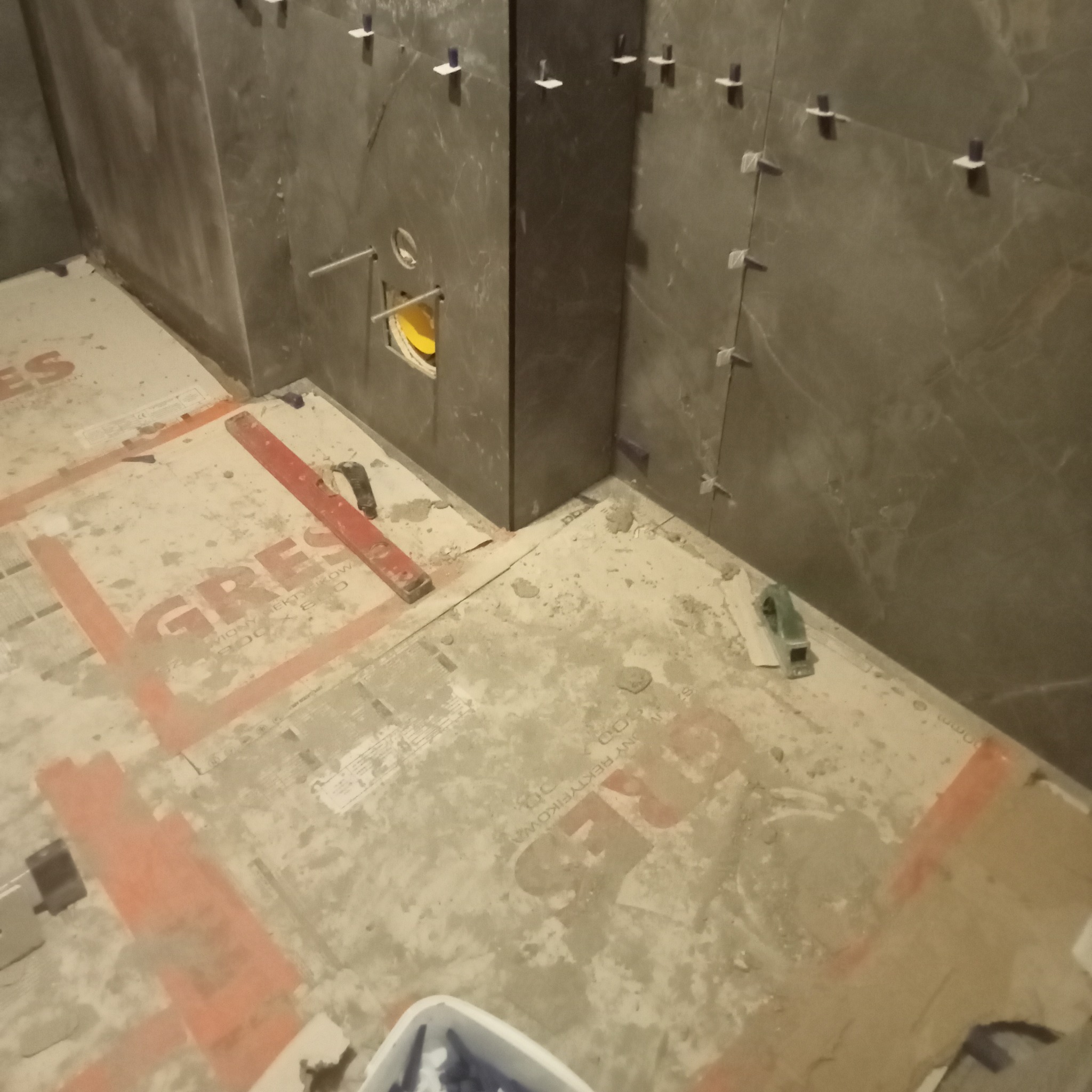 Układanie ciemnych, marmurkowych płytek w łazience. Widoczne krzyżyki dystansowe, poziomica i przygotowane podłoże. W tle otwór instalacyjny.
