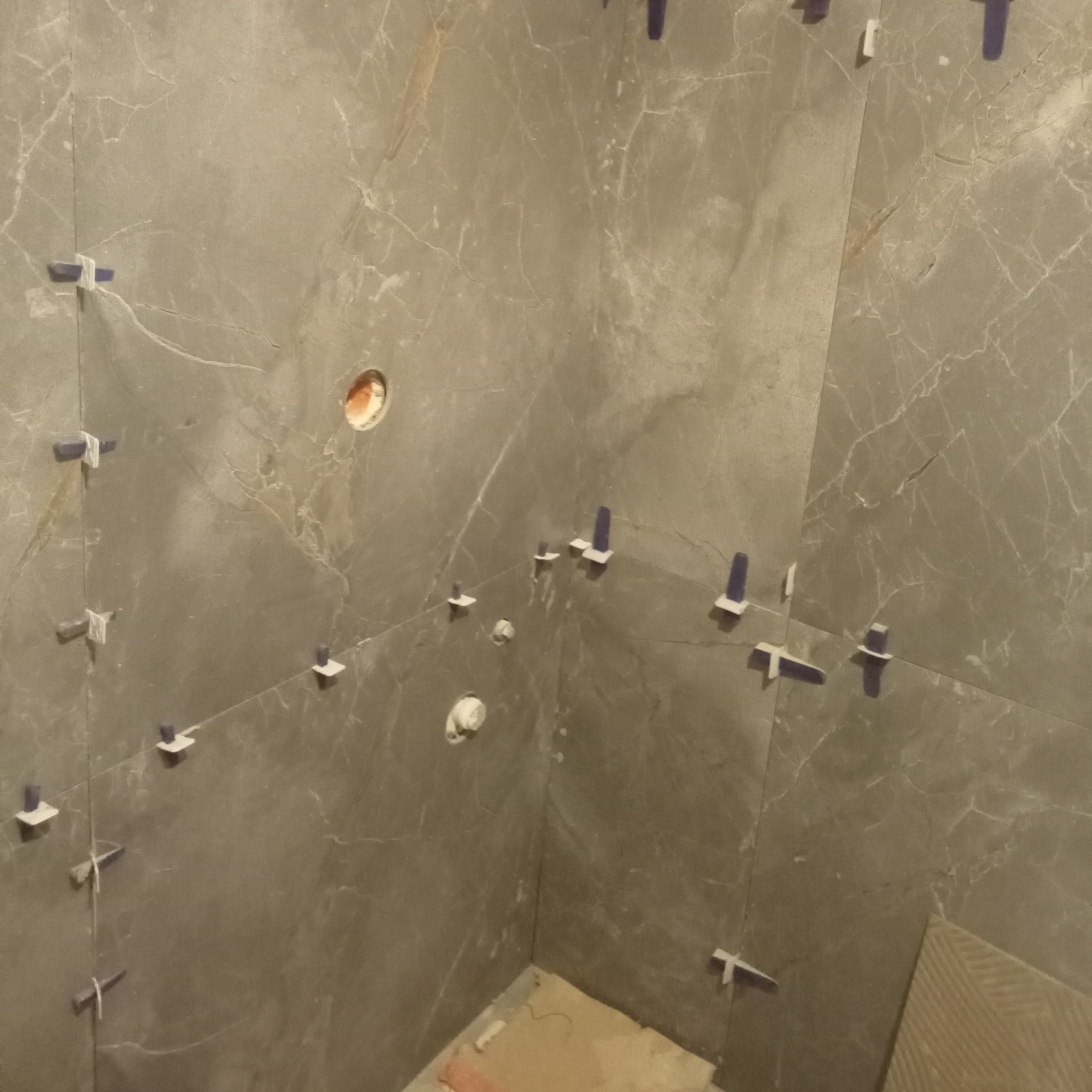 Szare płytki imitujące marmur, ułożone na ścianach łazienki, z widocznymi elementami systemu poziomowania płytek i otworami na instalacje.