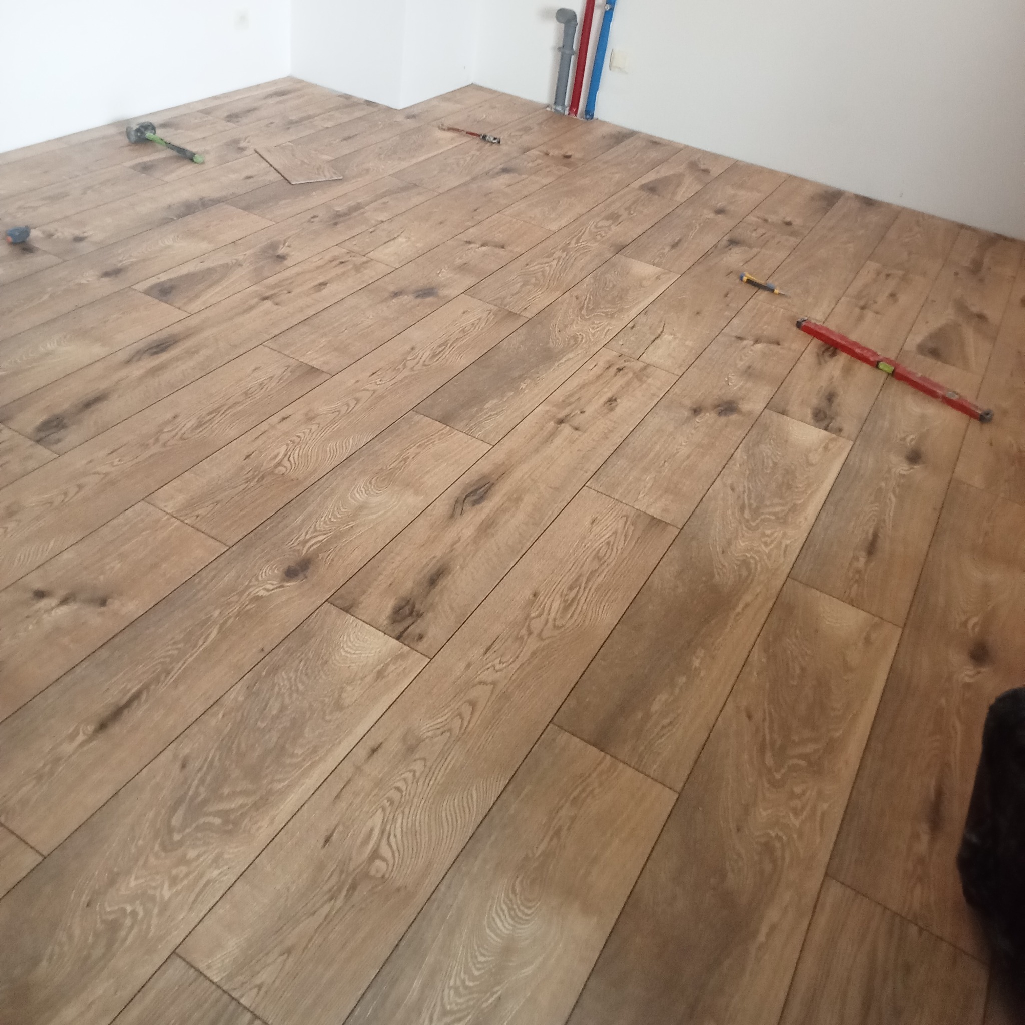 Układanie paneli podłogowych w jasnym odcieniu drewna. Widoczne narzędzia: poziomica, młotek gumowy i ołówek, co sugeruje etap montażu. Pionowe rury w tle.