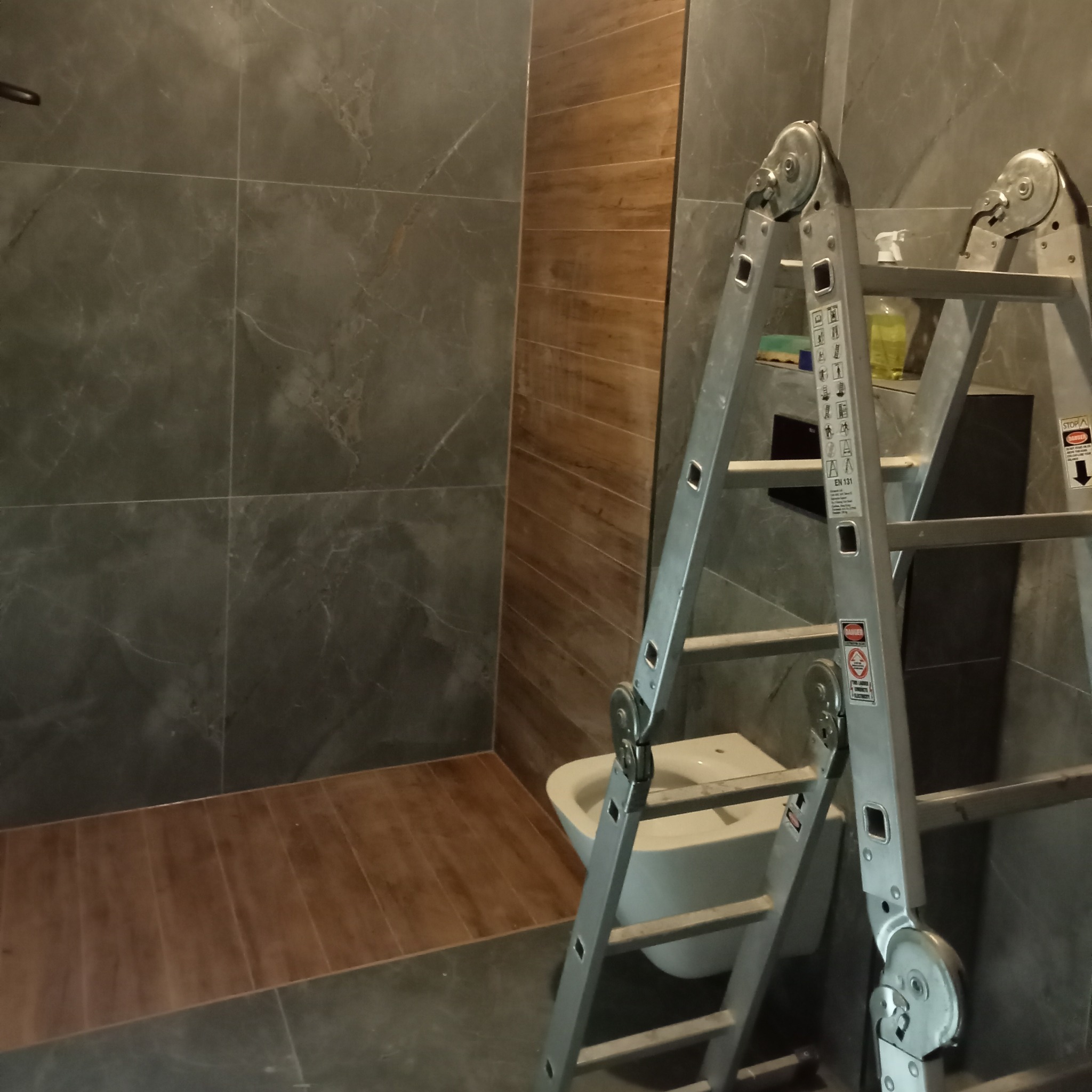 Wykończona łazienka z szarymi płytkami imitującymi marmur i drewnopodobnymi na podłodze. Widoczna biała toaleta i rozłożona aluminiowa drabina.