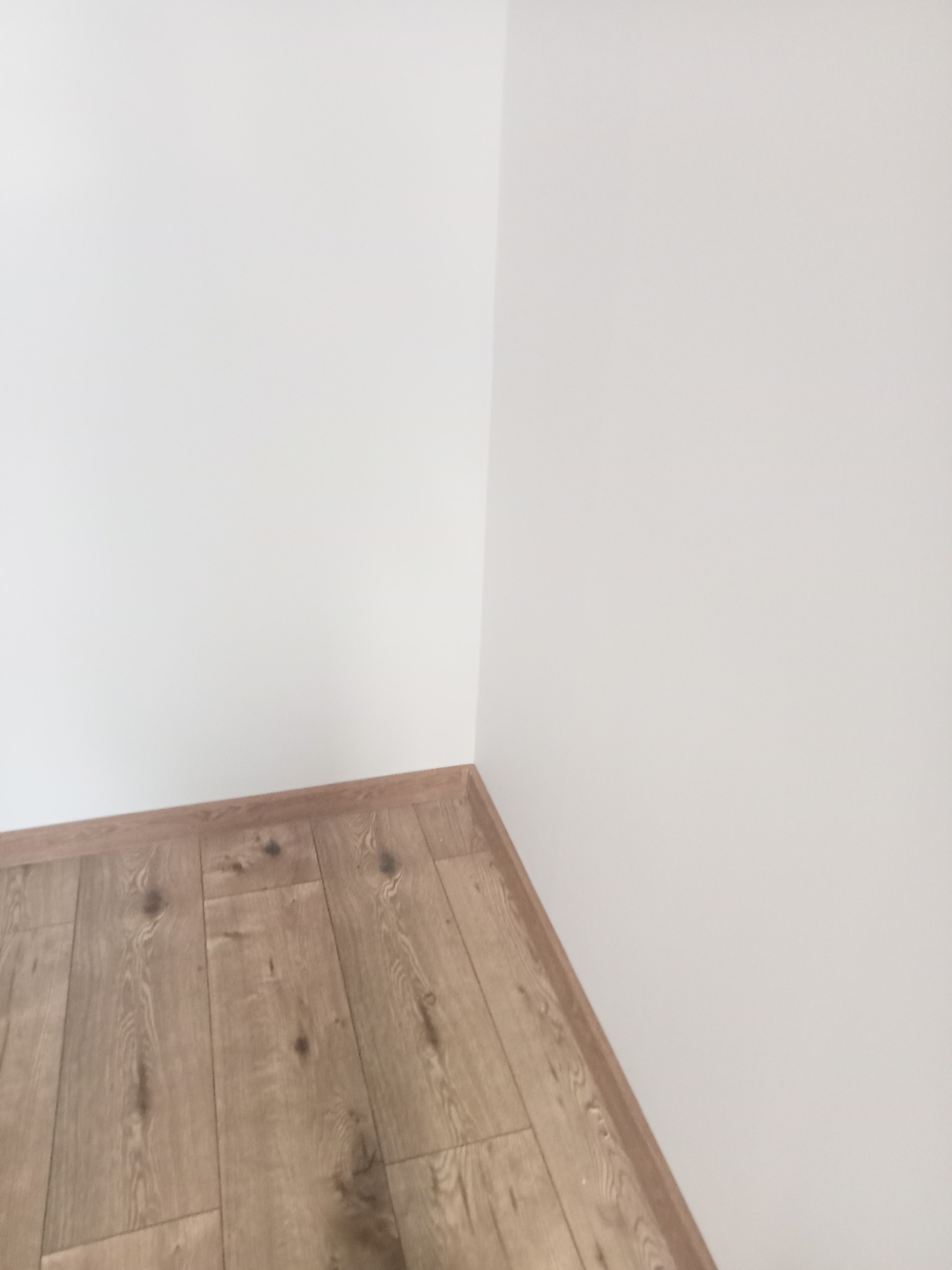 Jasne panele podłogowe ułożone w rogu pomieszczenia z białymi ścianami. Widoczne listwy przypodłogowe w kolorze paneli.