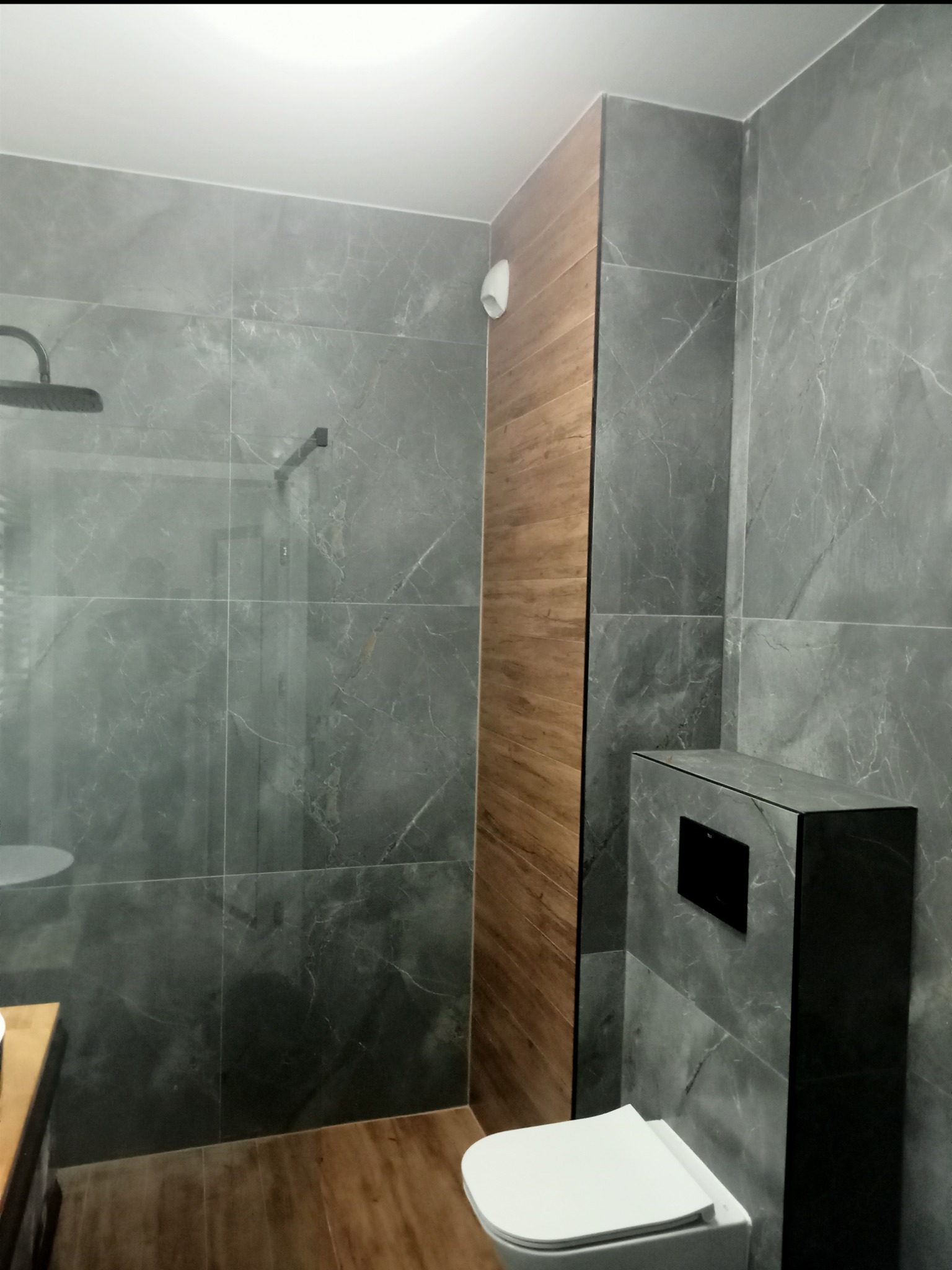 Nowoczesna łazienka z szarymi płytkami imitującymi marmur, drewnianym panelem ściennym i podwieszaną toaletą. Widoczna kabina prysznicowa typu walk-in.