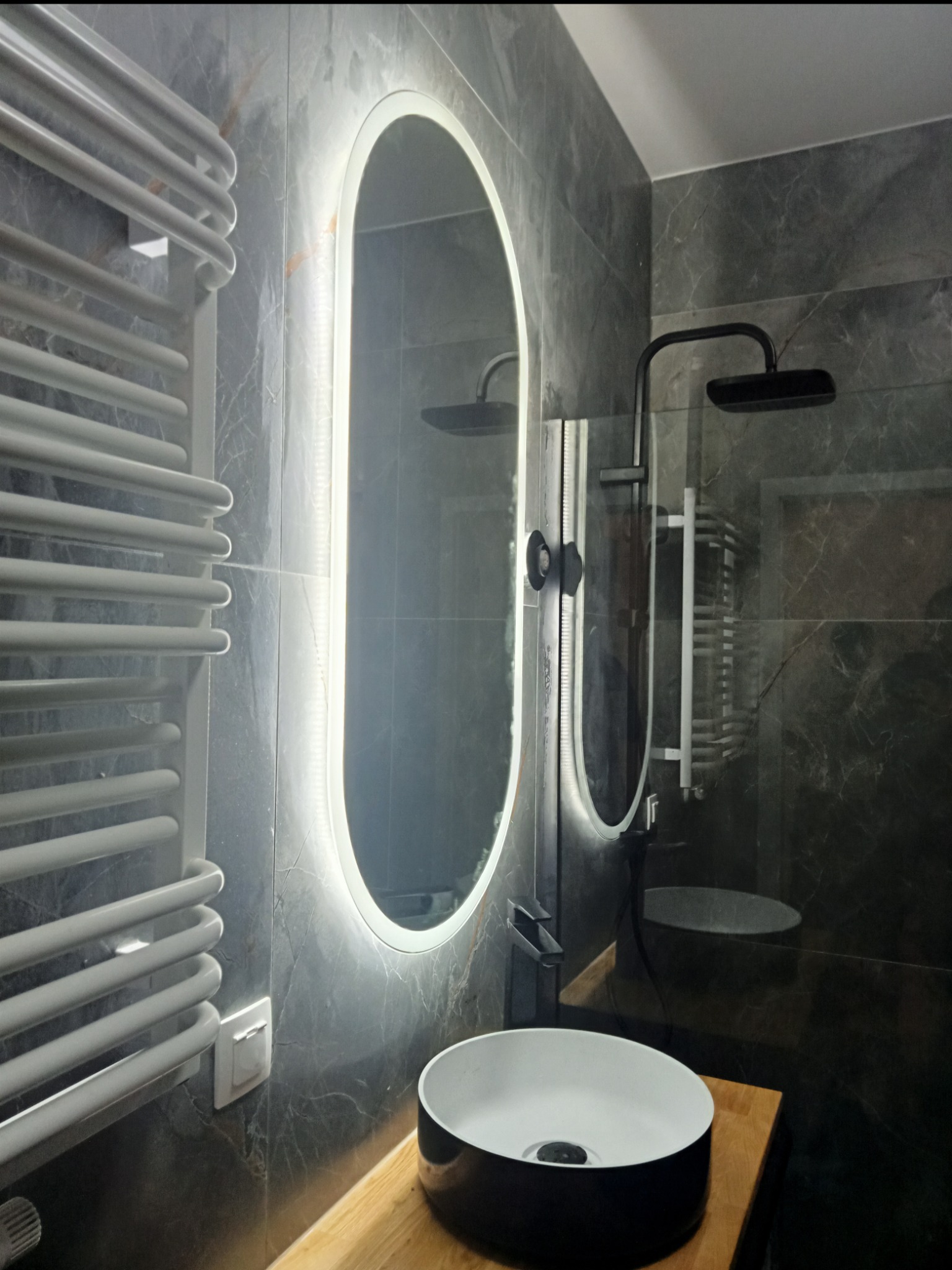 Nowoczesna łazienka z marmurowymi płytkami, podświetlanym lustrem i biało-czarną umywalką na drewnianym blacie. Grzejnik drabinkowy i czarna armatura dodają elegancji.