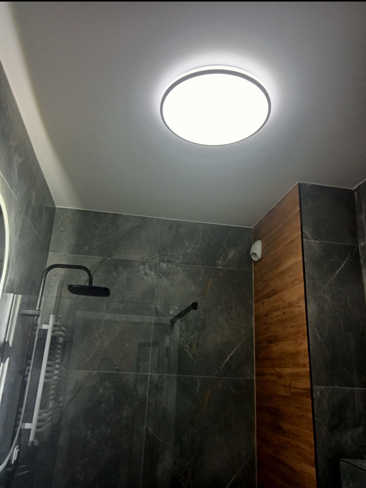 Nowoczesna łazienka po remoncie: ciemne płytki imitujące marmur, drewniane panele ścienne, okrągła lampa sufitowa i czarny zestaw prysznicowy.