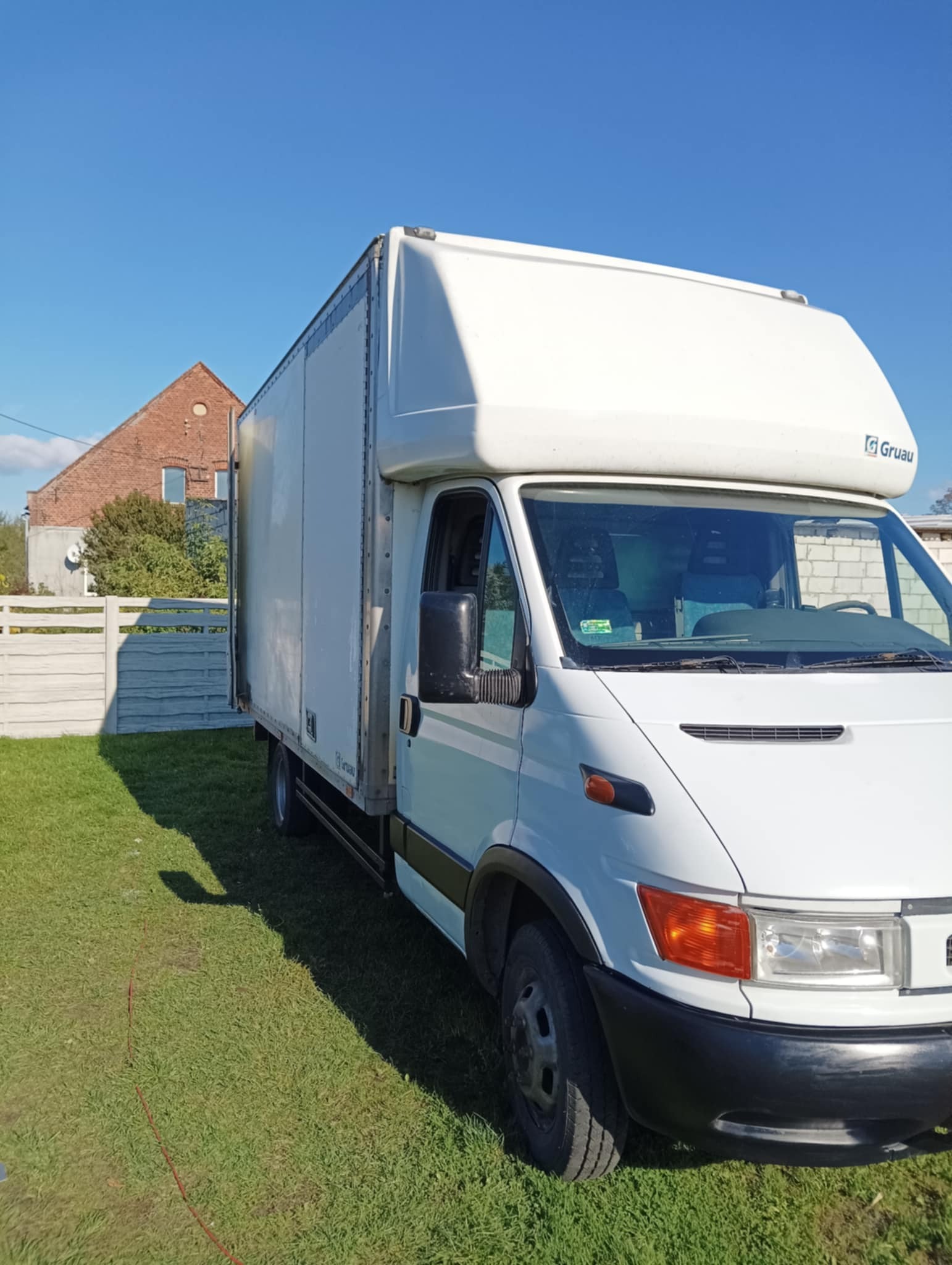 Biały samochód dostawczy z zabudową kontenerową marki Gruau, zaparkowany na trawie przed budynkiem z czerwonej cegły i białym płotem.