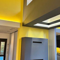 FlexiWorks-Przemysław Wolnik - Nowoczesne wnętrze z żółtymi ścianami, szarym kominkiem i podświetlanym sufitem kasetonowym, widok na przedpokój z czarnymi drzwiami i krzyżem na ścianie.