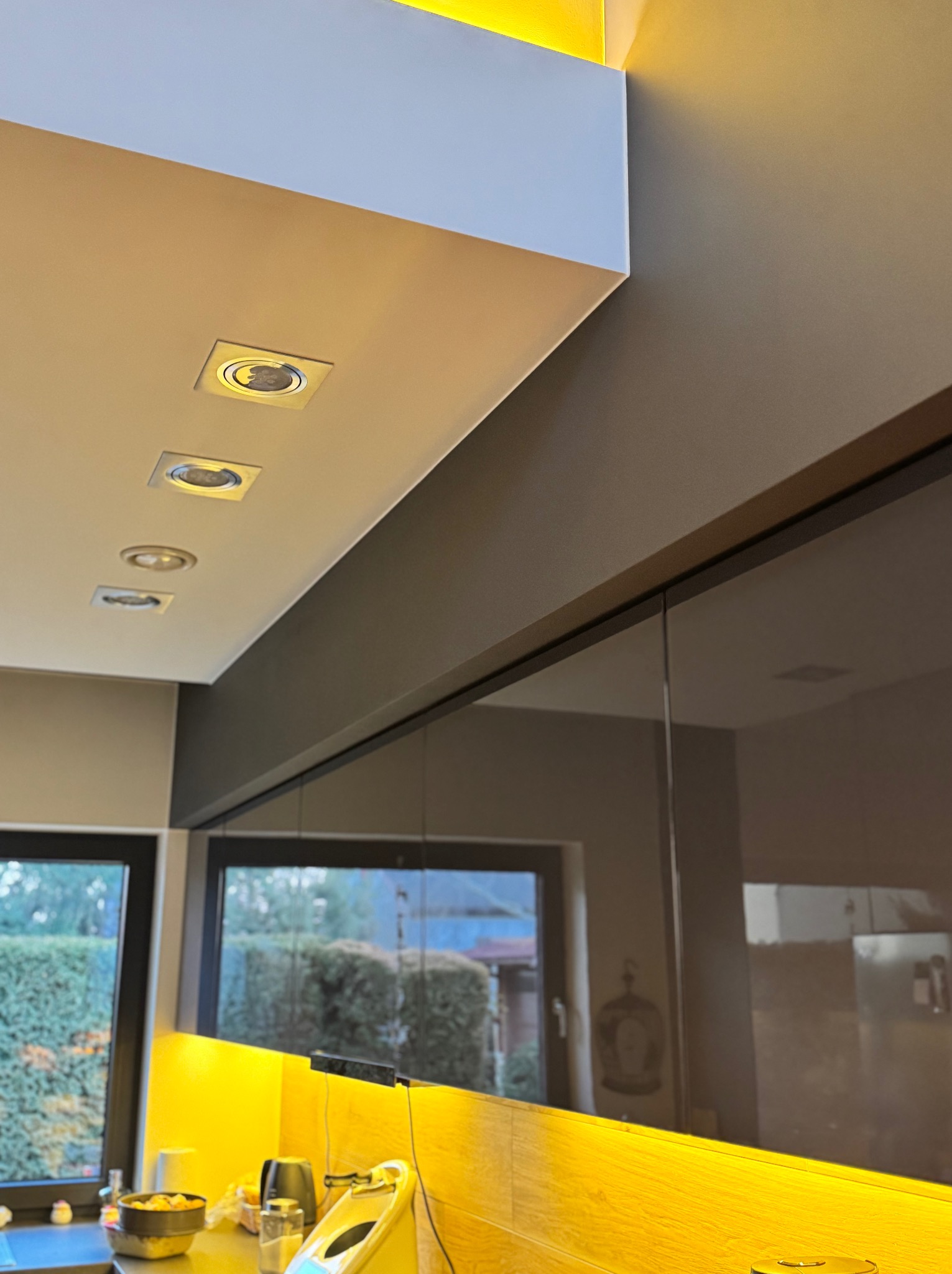 Nowoczesna kuchnia z oświetleniem punktowym LED w suficie, szarymi ścianami i żółtymi elementami zabudowy, widok na okno z zielenią na zewnątrz.