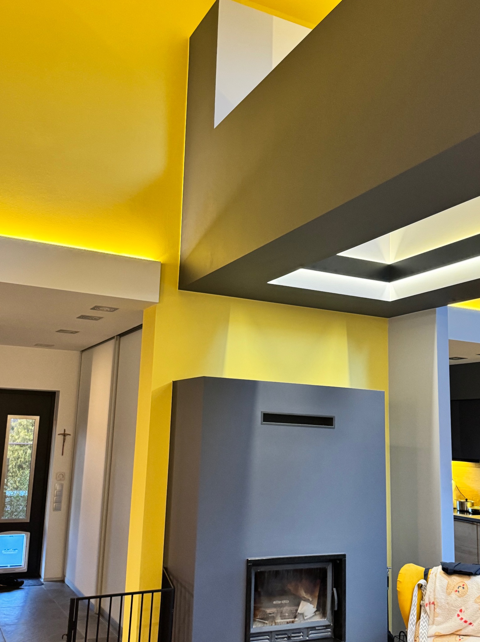 Nowoczesne wnętrze z żółtymi ścianami, szarym kominkiem i podświetlanym sufitem kasetonowym, widok na przedpokój z czarnymi drzwiami i krzyżem na ścianie.