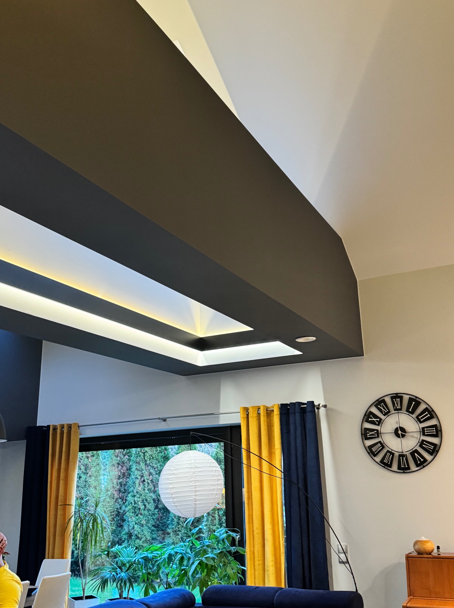 Nowoczesny sufit z podświetleniem LED w salonie, z widocznymi elementami dekoracyjnymi jak zasłony w kolorze granatowym i żółtym oraz zegar ścienny w stylu retro.