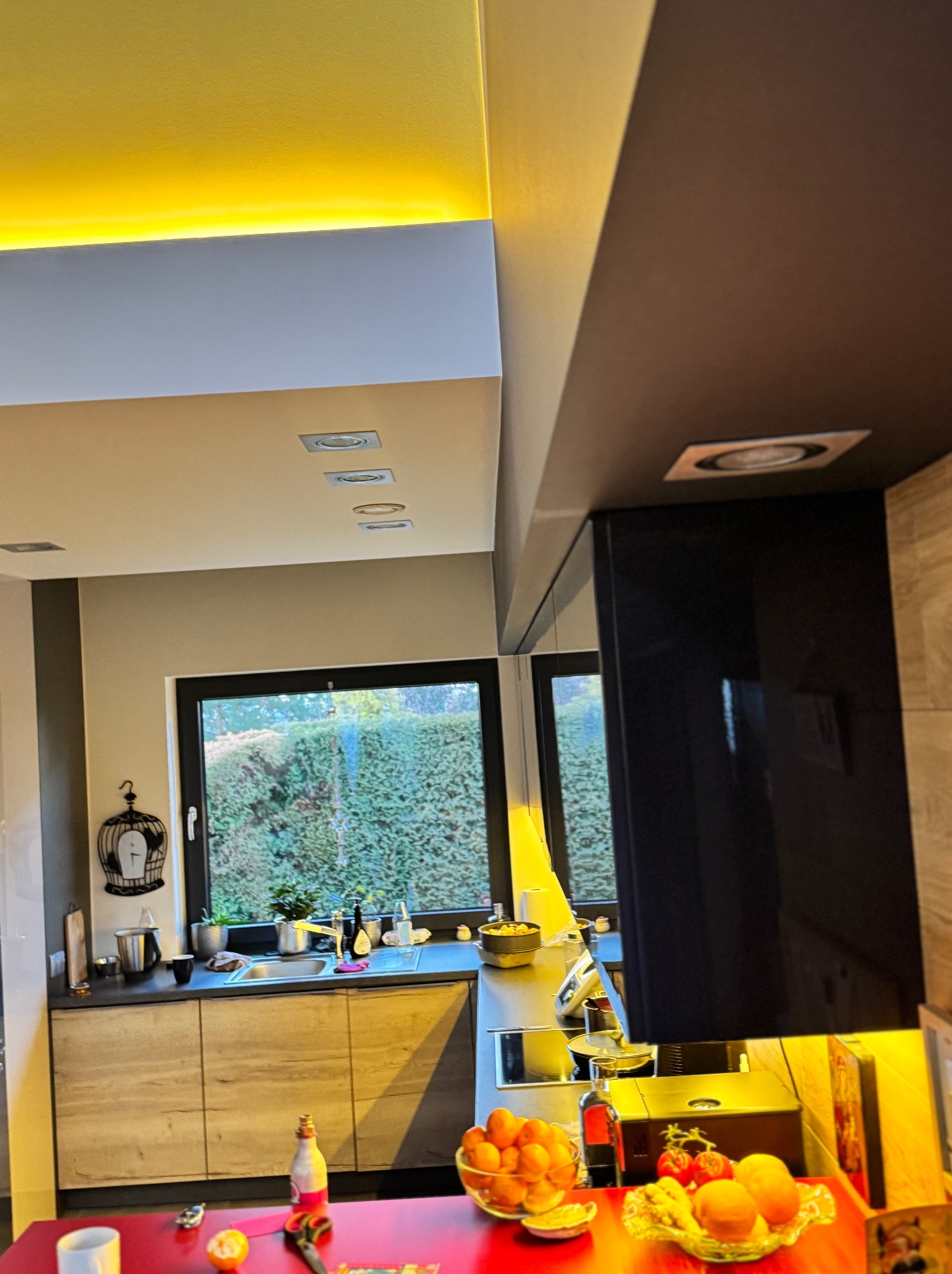 Nowoczesna kuchnia z podświetlanym sufitem, widok na okno z zielenią, czarne szafki i drewniane fronty, owoce na stole.