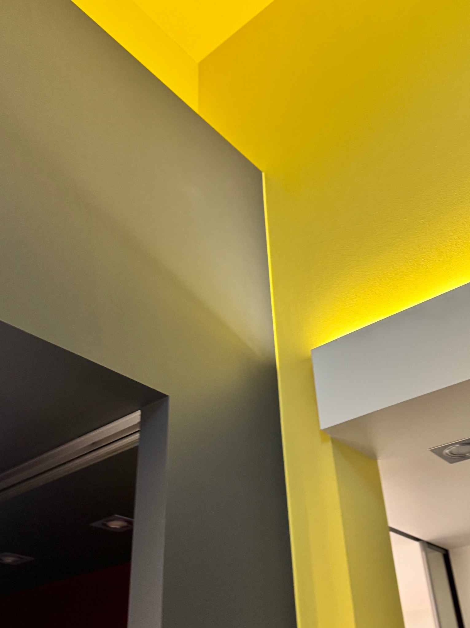 Fragment wnętrza z pomalowanymi ścianami w odcieniach szarości i intensywnej żółci, z oświetleniem LED w suficie podwieszanym, widok z dołu.