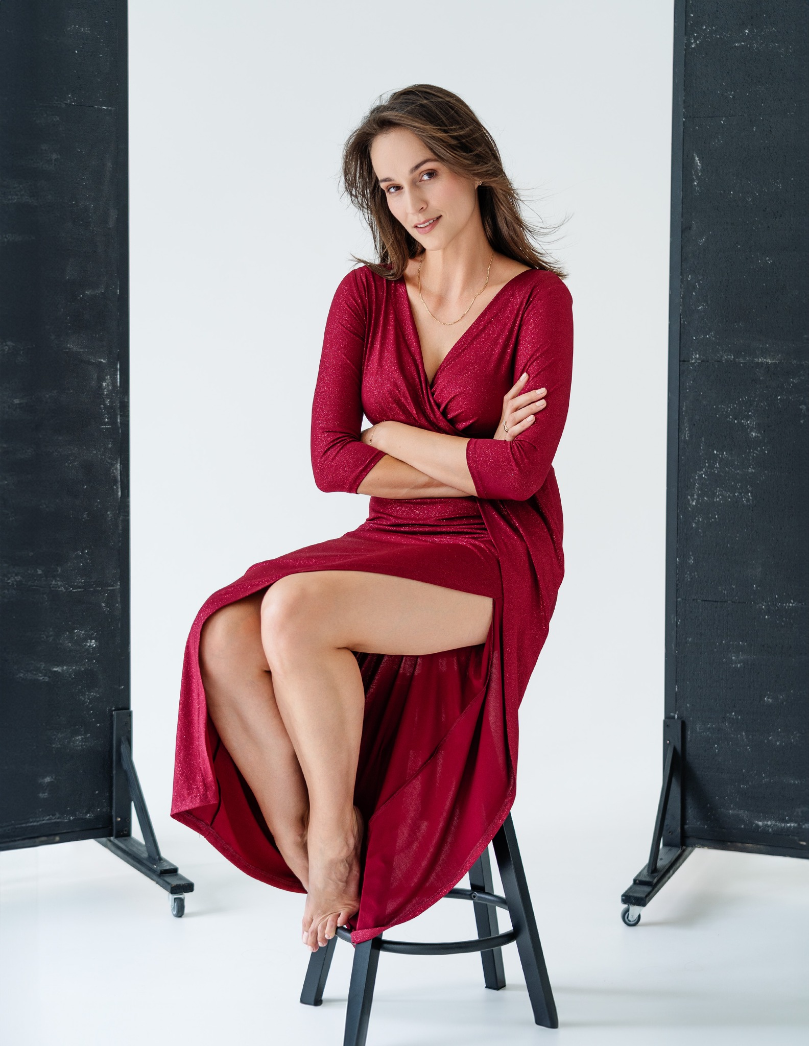Kobieta w bordowej, błyszczącej sukience z rozcięciem, siedząca na czarnym stołku na tle białej ściany z czarnymi panelami po bokach, pozująca z założonymi rękami.