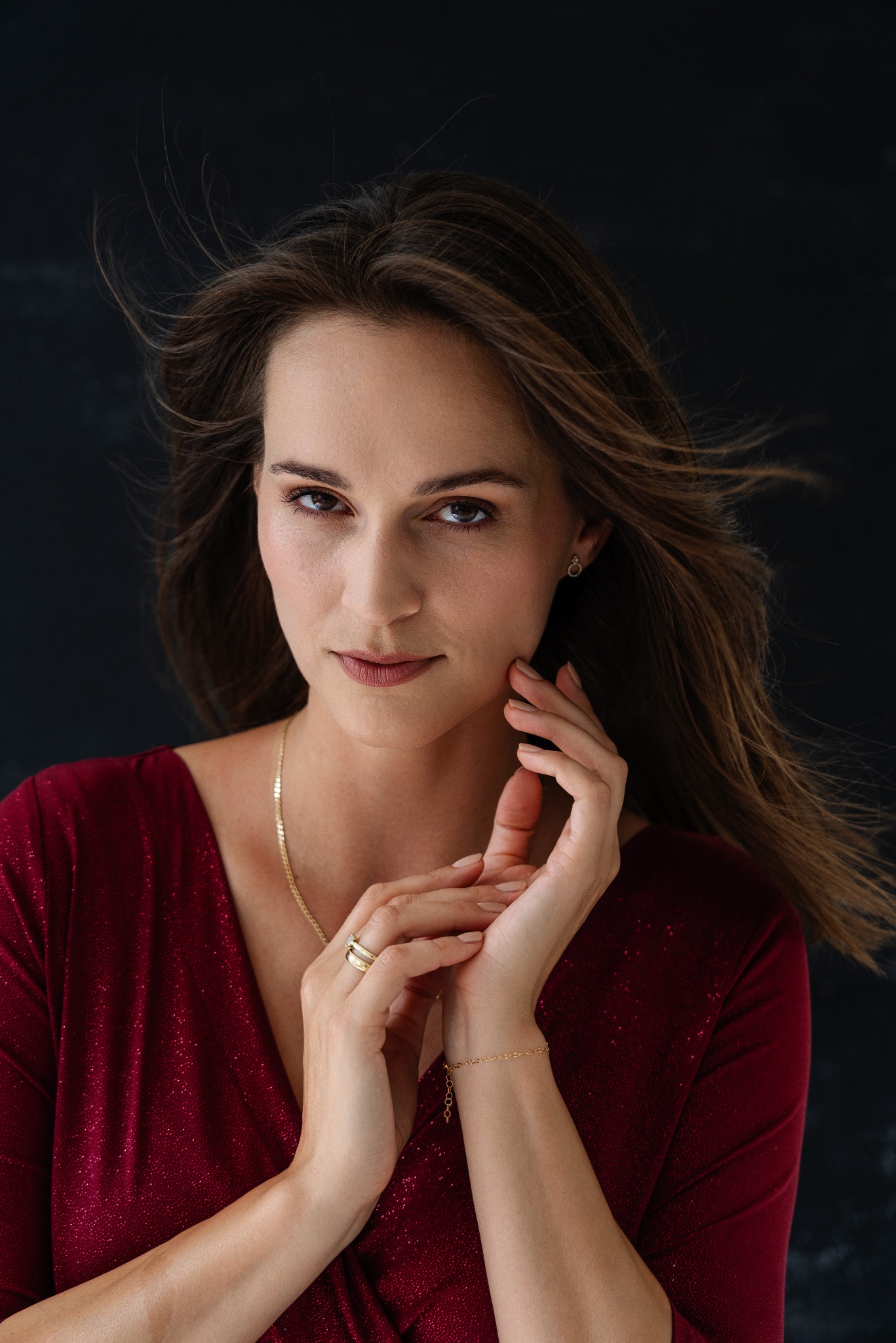 Portret kobiety w bordowej, błyszczącej sukience, z rozwianymi włosami i delikatnym makijażem, pozującej na ciemnym tle. Widoczne złote dodatki: kolczyki, naszyjnik, bransoletka i pierścionek.