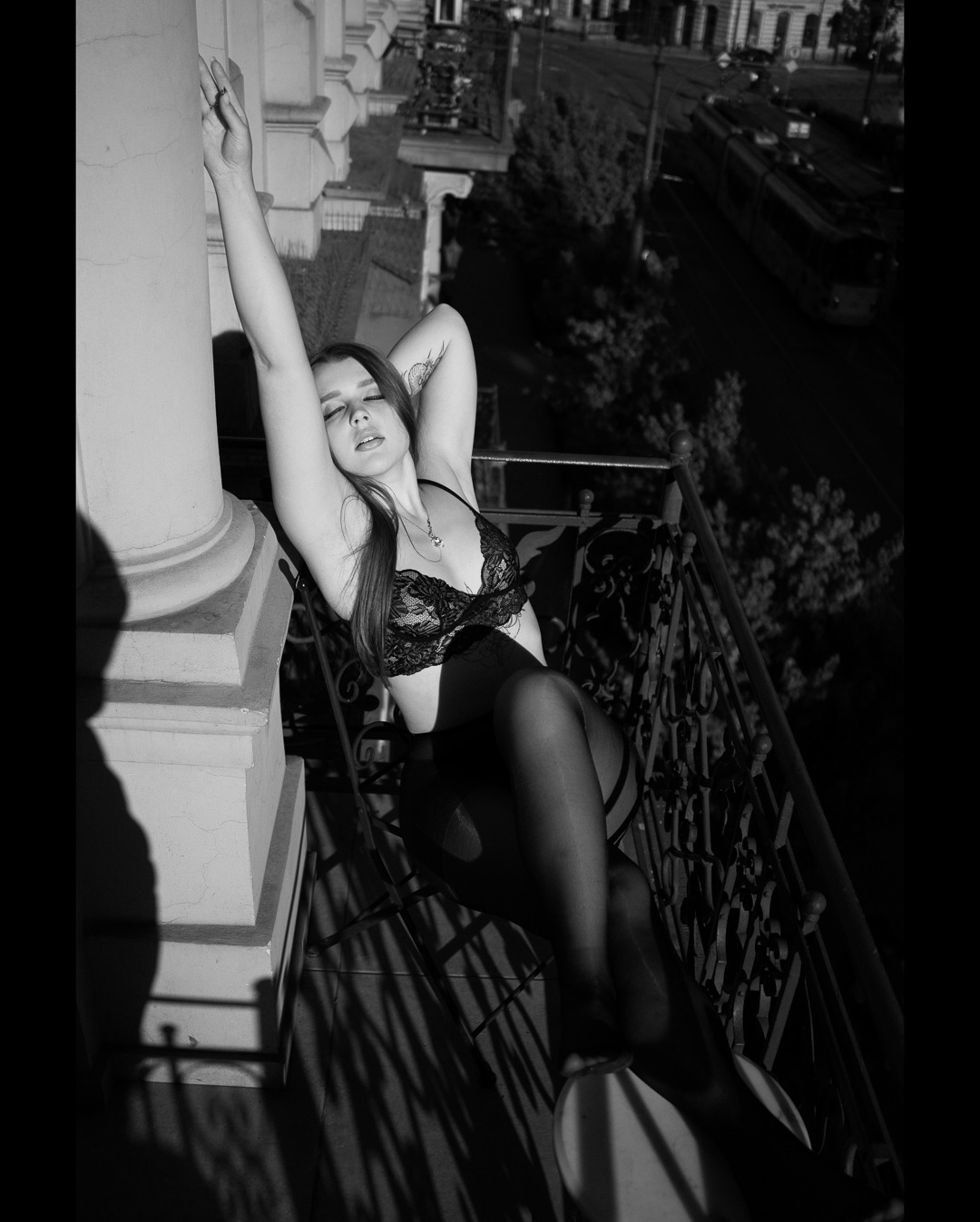 Czarno-białe zdjęcie kobiety w bieliźnie, opierającej się o kolumnę na balkonie z widokiem na ulicę z tramwajem w tle.