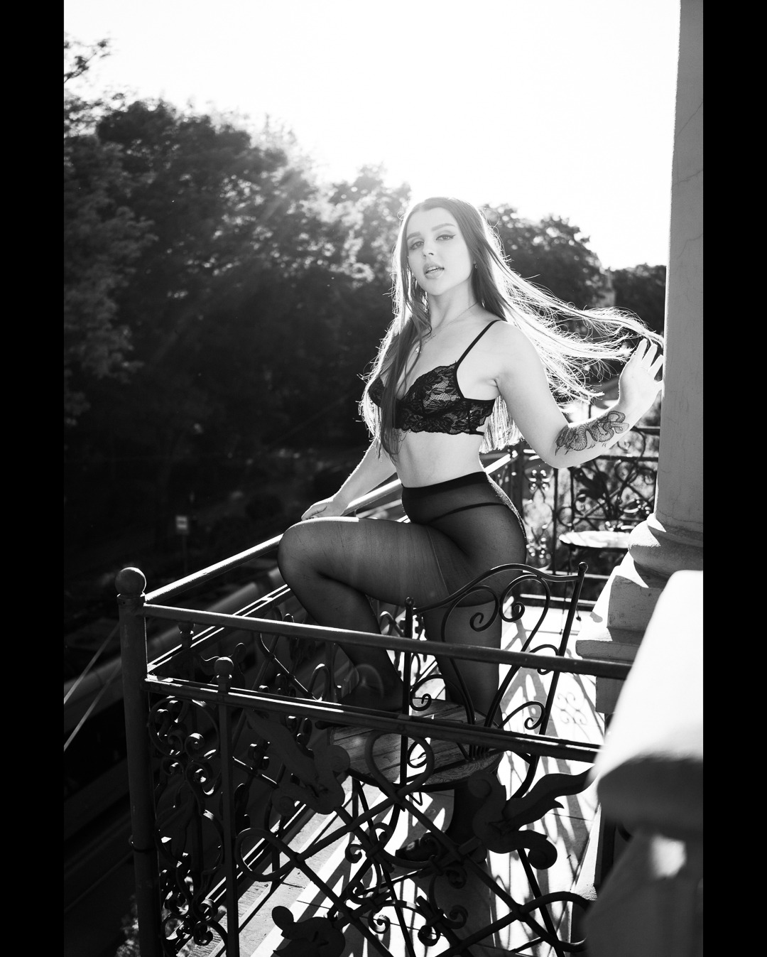 Czarno-białe zdjęcie kobiety w bieliźnie na balkonie z ozdobną balustradą, włosy rozwiane na wietrze, tatuaż na ramieniu, widok na zieleń w tle.