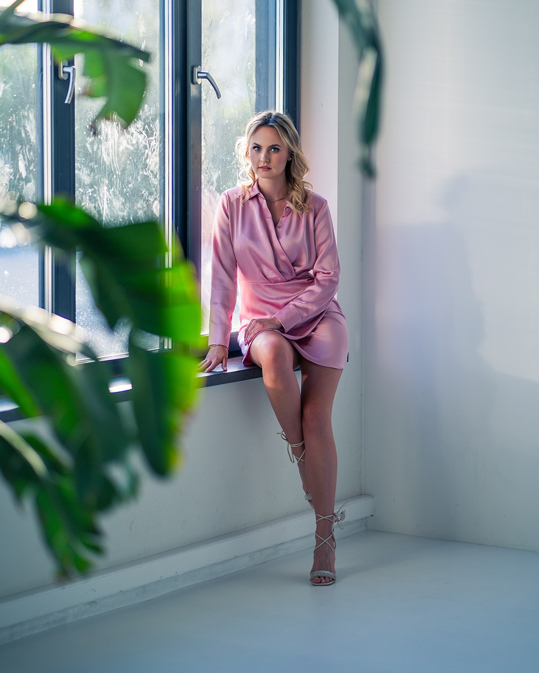 Kobieta w różowej sukience siedzi na parapecie okna z zielonymi roślinami w kadrze, patrząc w obiektyw.