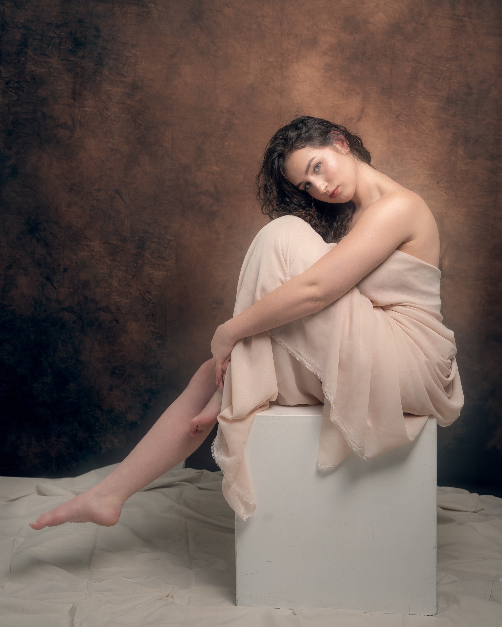 Portret kobiety z kręconymi włosami, owiniętej w beżową tkaninę, siedzącej na białym sześcianie przed brązowym, teksturowanym tłem.